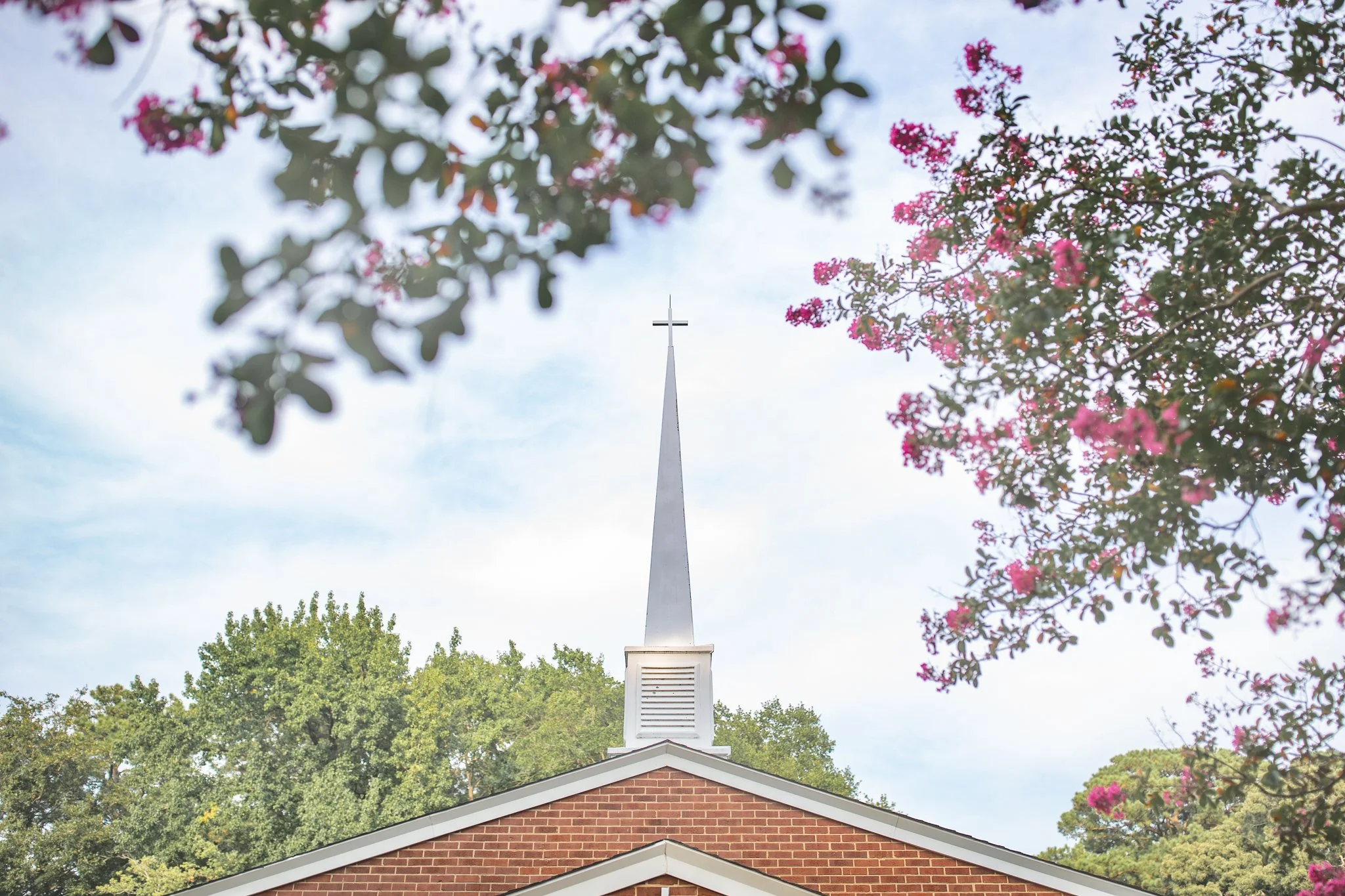 View of a church steeple with a cross on top, seen from below through blooming pink flowers and green trees on a partly cloudy day.