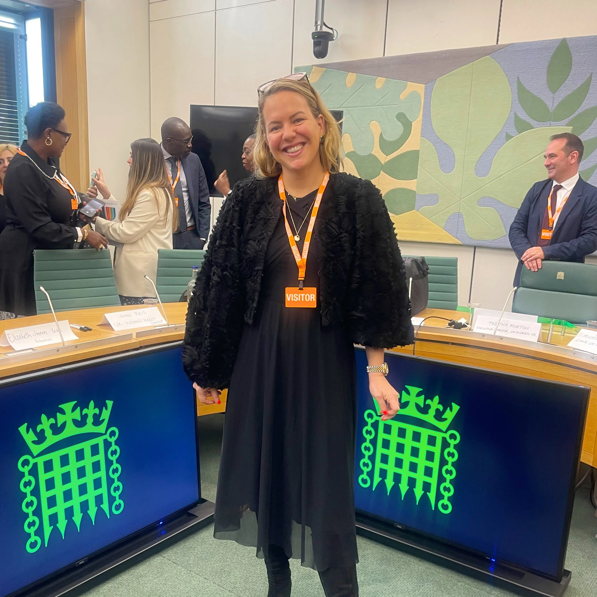 A woman with blonde hair smiling and wearing a visitor badge stands in front of a decorated backdrop at the UK Parliament. In the background, there are other people engaging in conversation, some wearing badges.