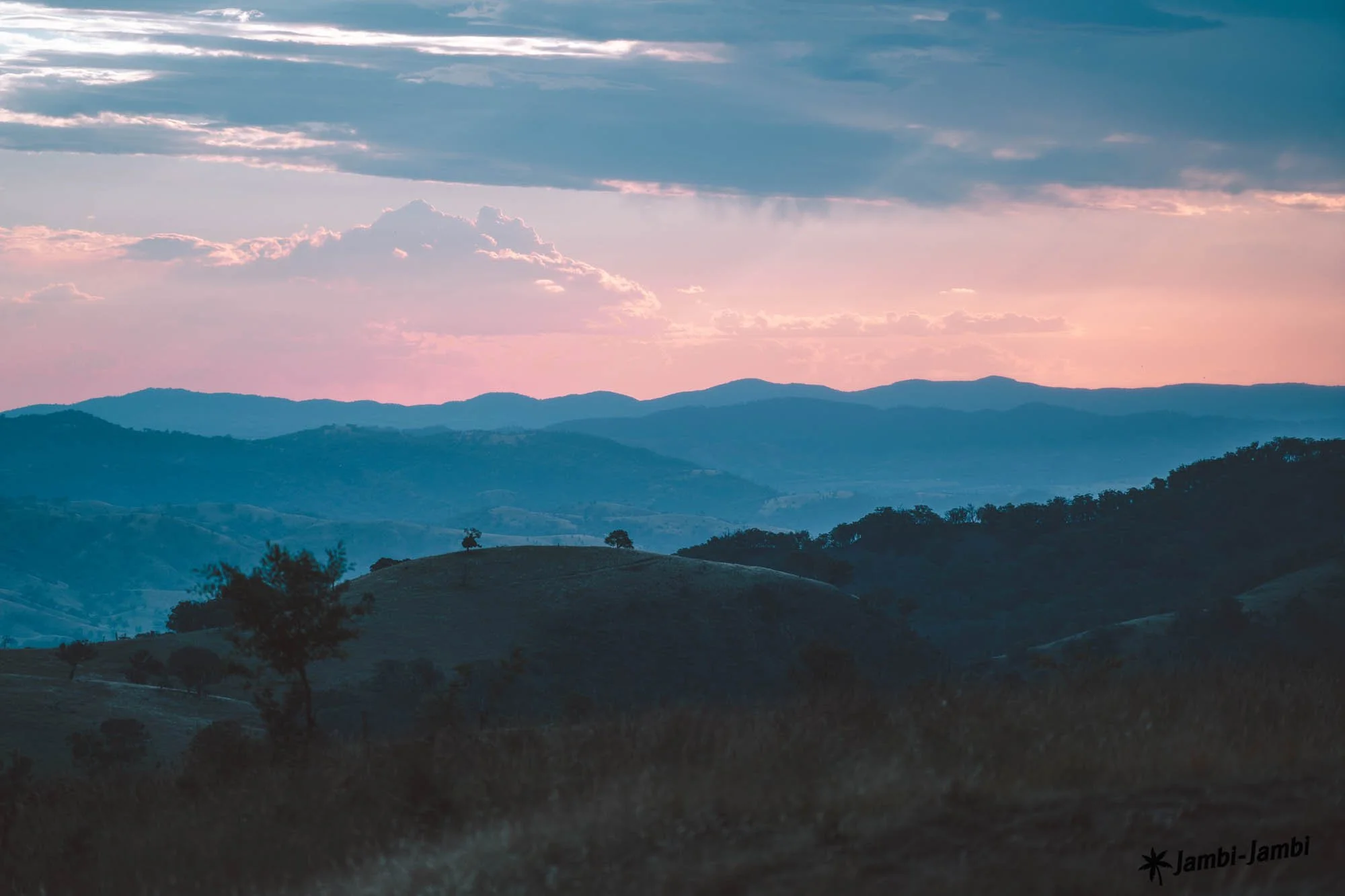 A scenic view of rolling hills and mountains during dusk, with a colorful sky displaying pink and blue hues, scattered clouds, and calm natural landscape.