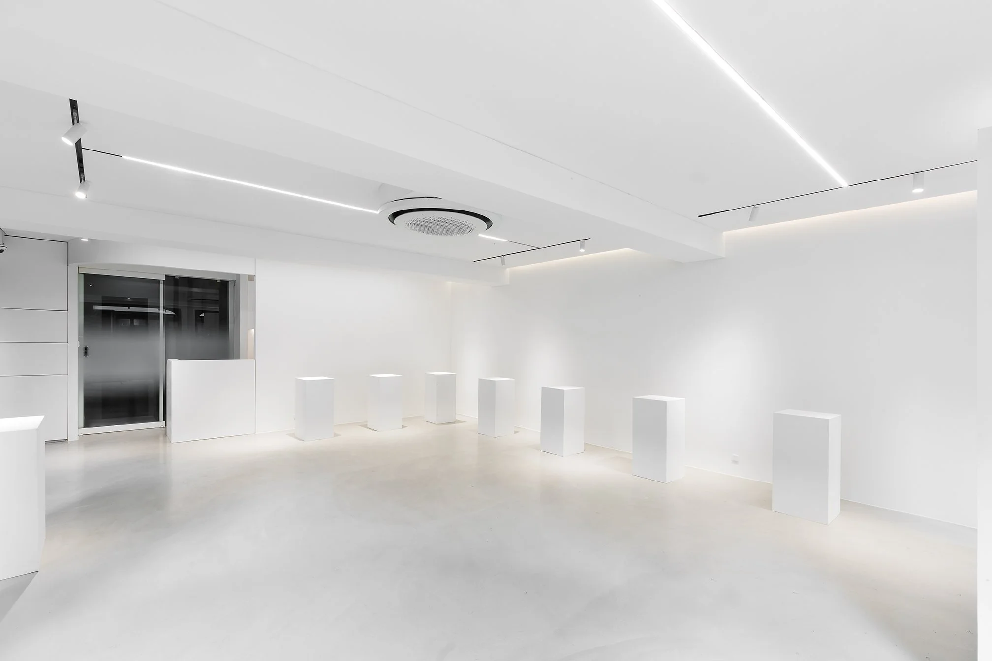 A minimalist, all-white room with geometric white pedestal stands and sleek ceiling lighting.