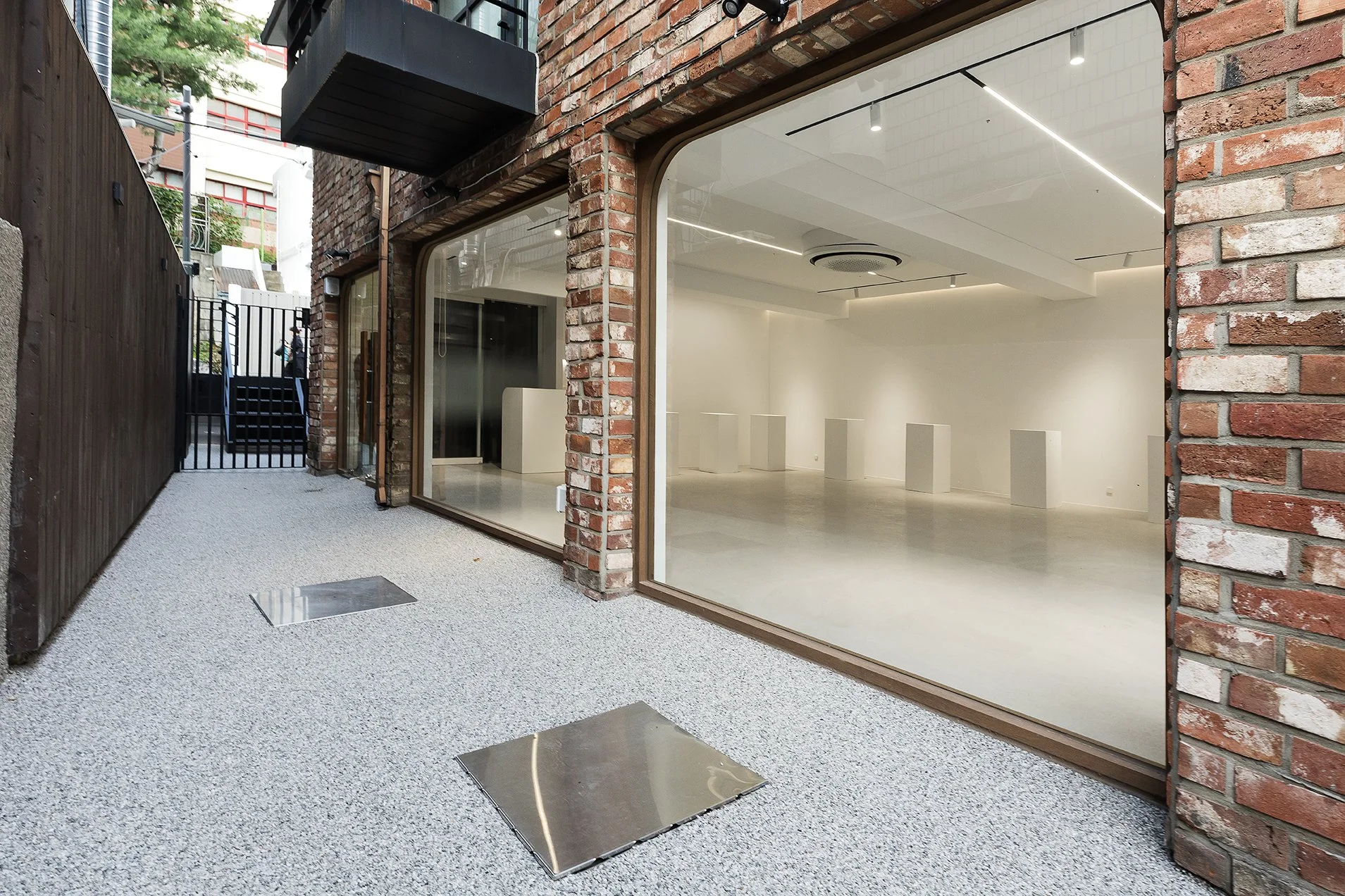 Photo of an empty art gallery with white walls, polished concrete floor, and display pedestals inside a brick building, viewed through a large window from an outdoor pathway with gravel flooring and a dark wooden fence.
