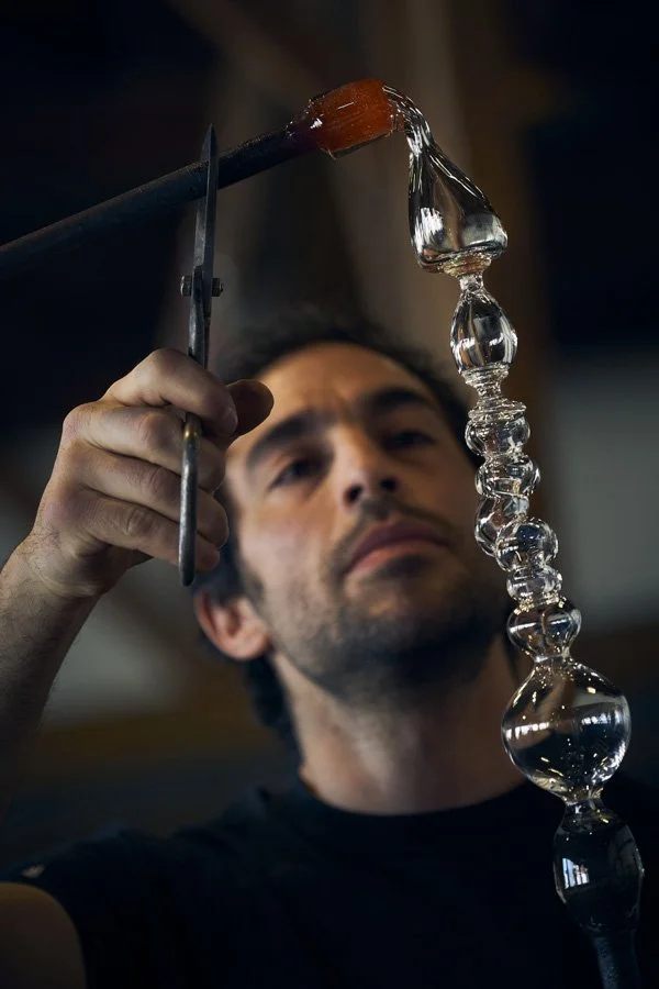 Glass artist shaping a glass sculpture using tools in a workshop.