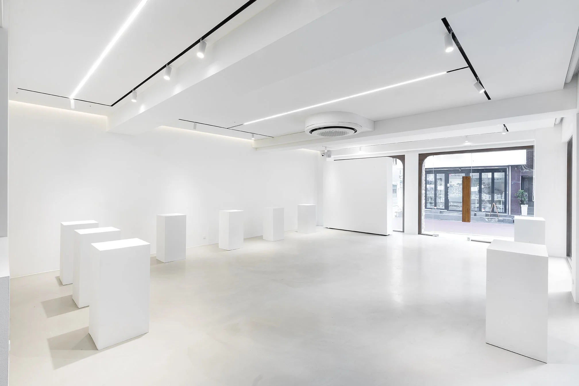 A bright, modern, art gallery with white walls, ceiling, and floor. Several white display pedestals are aligned near the front, and large windows allow natural light to fill the space. Minimalist ceiling lighting and a circular air vent are visible.