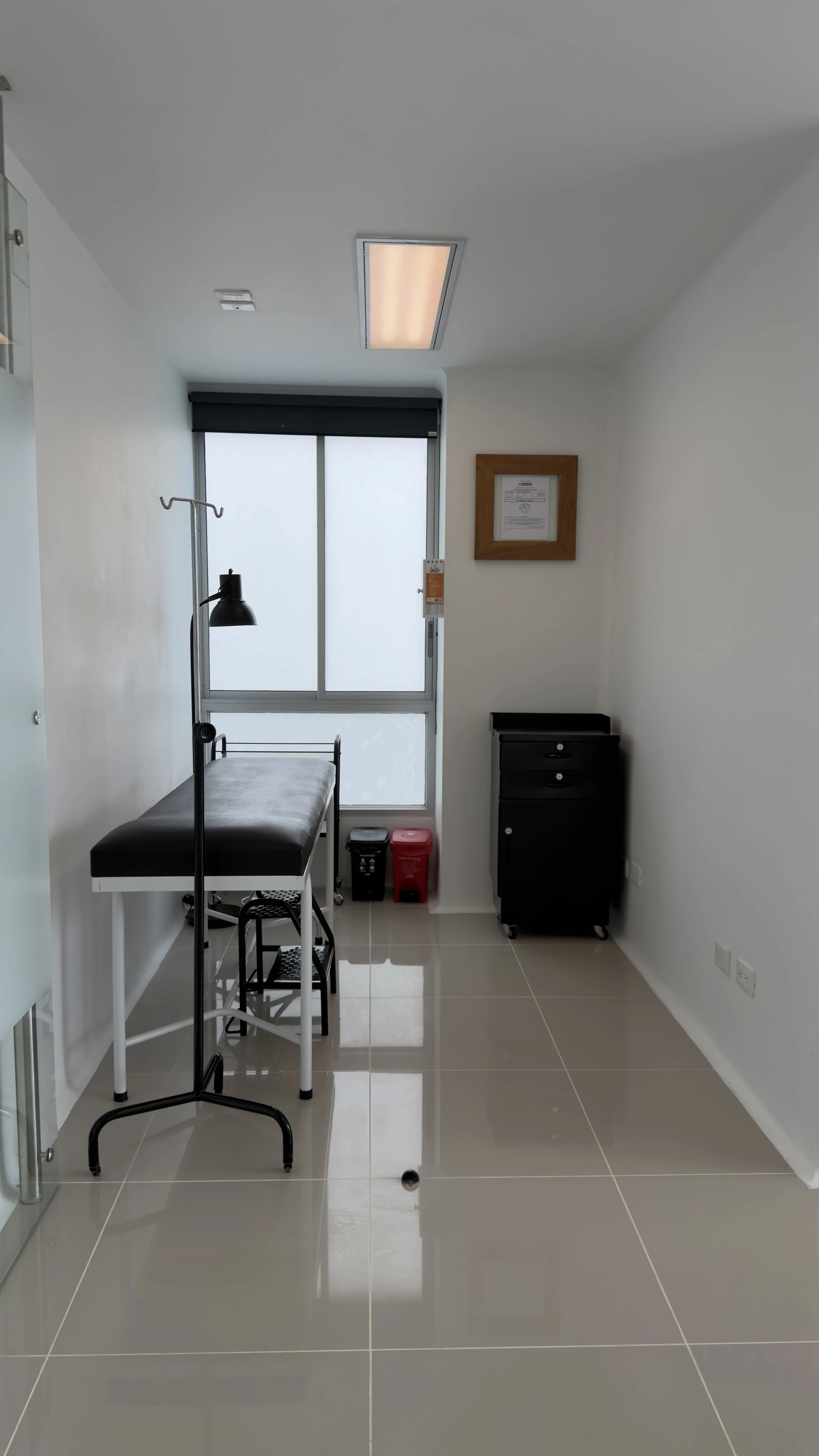Medical examination room with a black examination table, a tall medical lamp, a window with frosted glass, a framed certificate on the wall, and a small black cabinet.