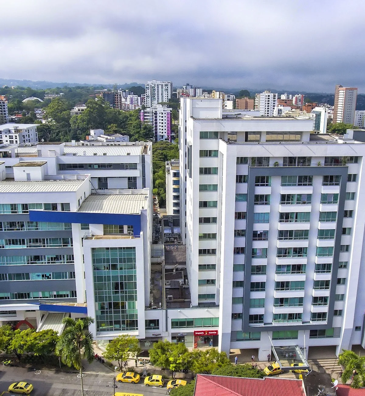 An aerial view of modern high-rise buildings in an urban area with a cloudy sky in the background, and a street with yellow taxis at the bottom.
