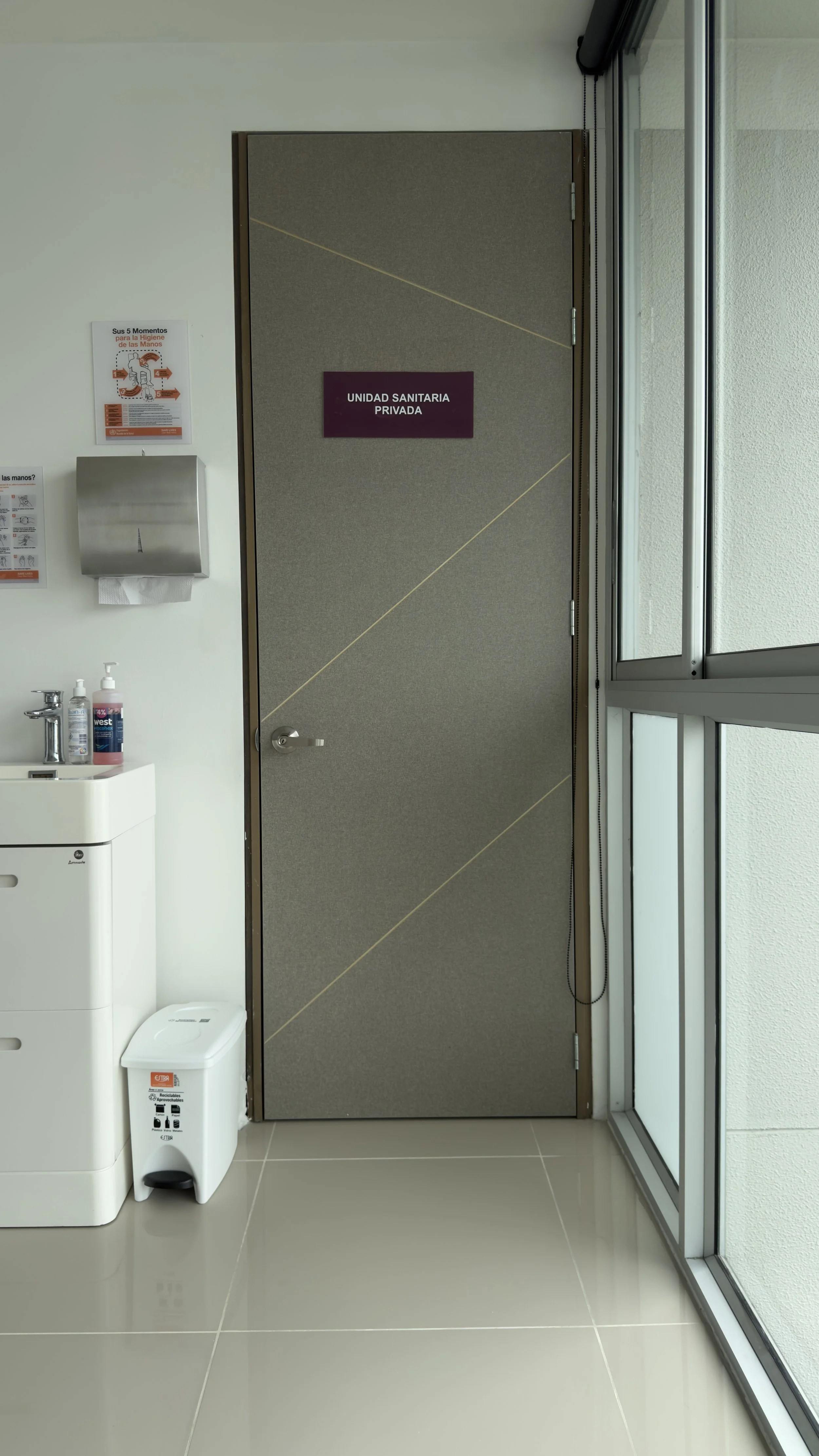Hospital hallway with a door labeled 'Unidad Sanitaria Privada', hand sanitizer dispenser, hand towels, and a trash bin, next to large glass windows.