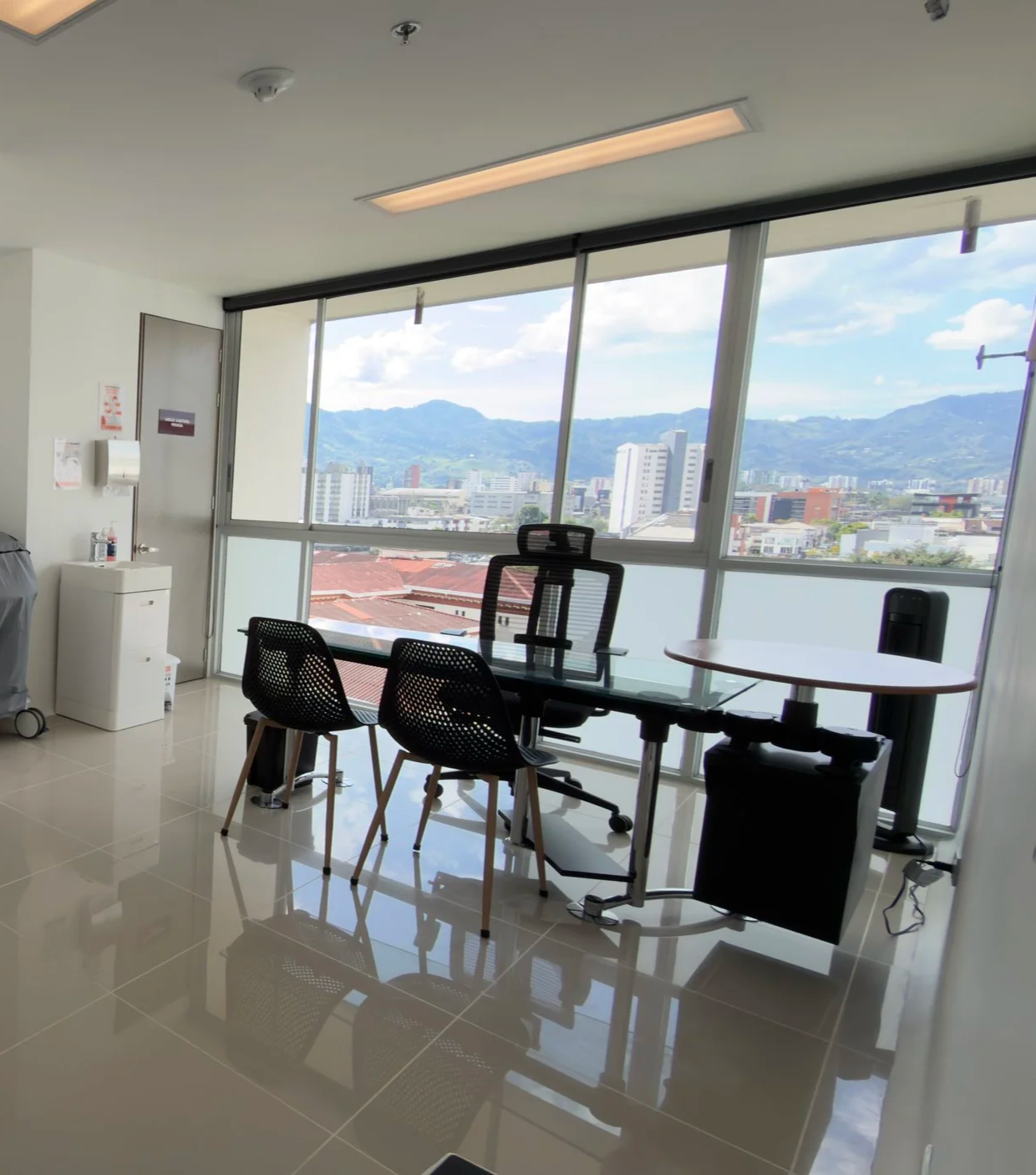 Office with large windows displaying a cityscape and mountains, black office chair, glass desk, two black chairs, and a small round table.