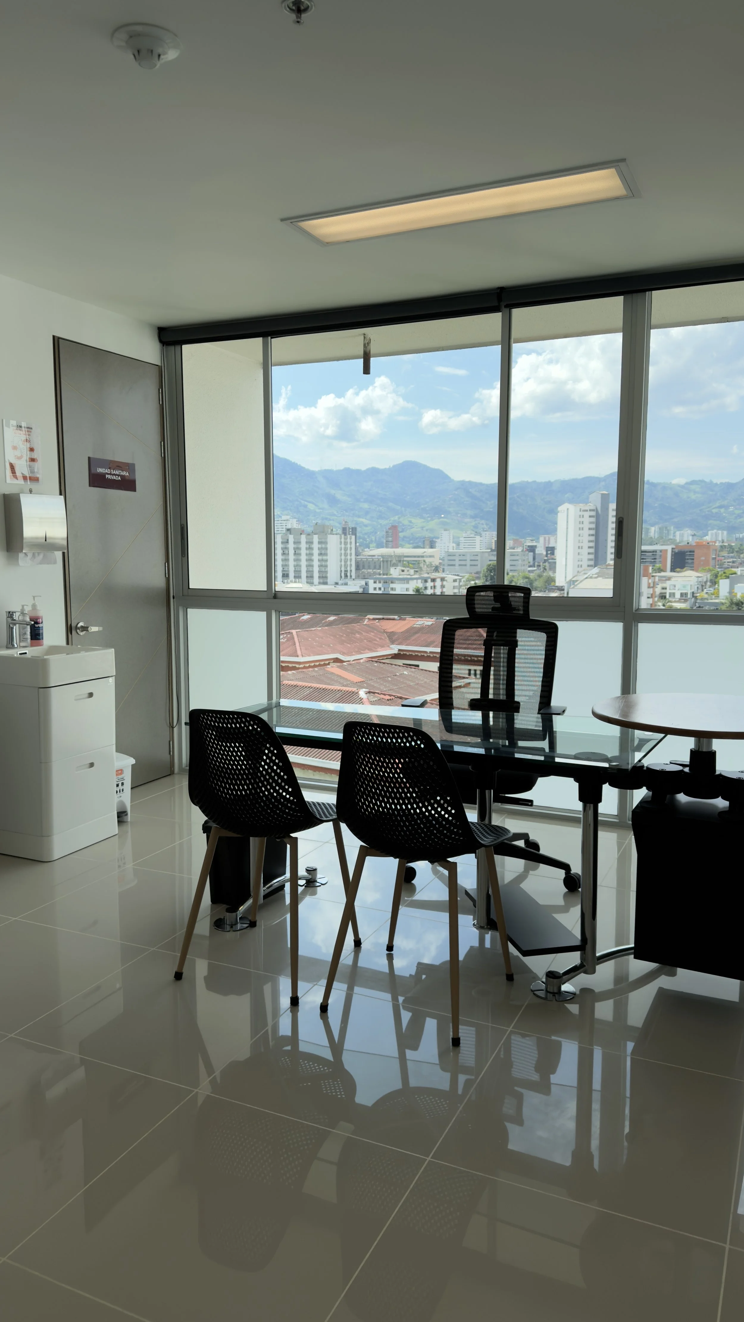 Office conference room with glass table, black chairs, window view of cityscape and mountains, ceiling light, water dispenser, and a door with sign.