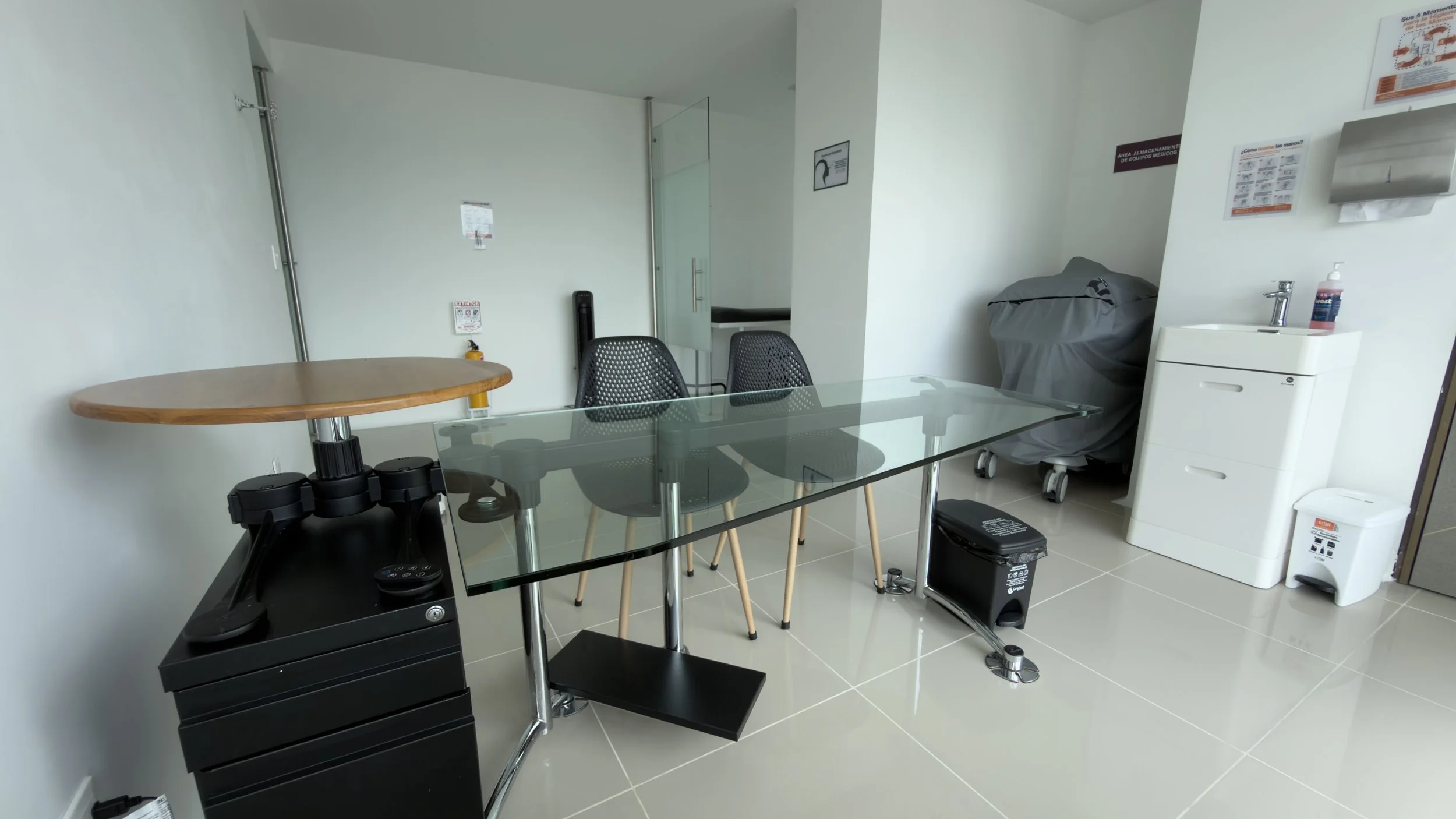 An office or medical consultation room with white walls and tiled flooring, featuring a glass-top table, black chairs, a small wooden table, a stationary scanner, a white cabinet, a water dispenser, a covered medical equipment cart, and various safet