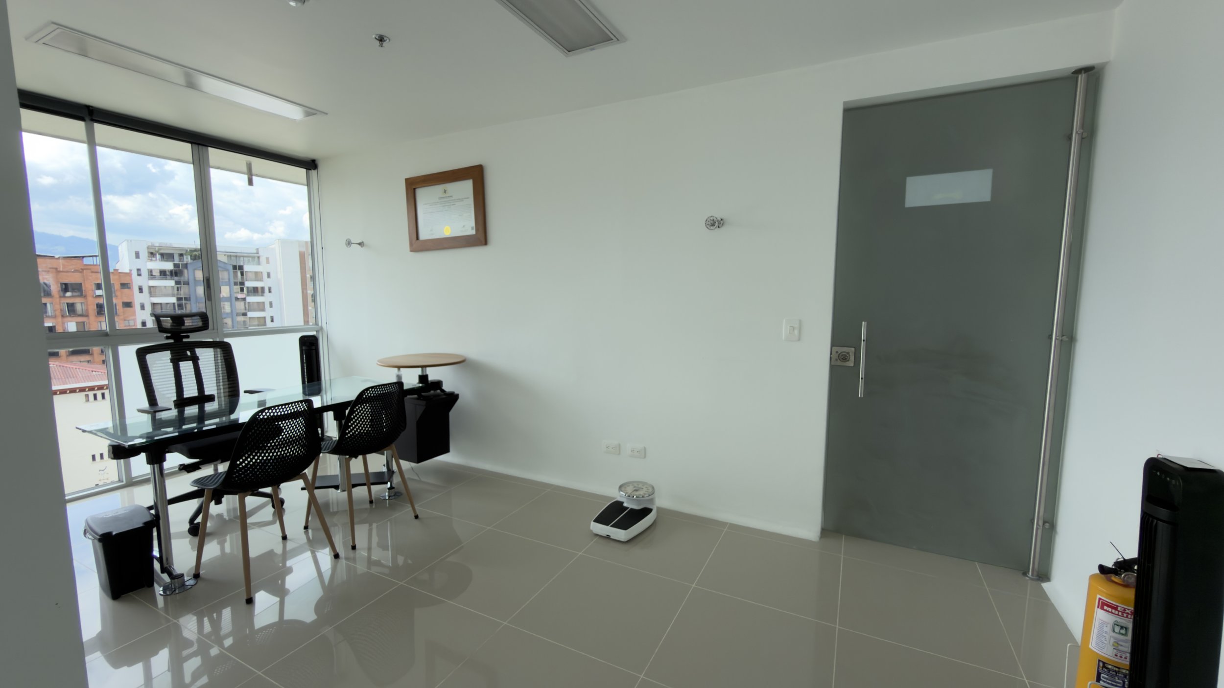 Modern office with a glass wall, black office chairs around a table, a framed certificate on the white wall, a spray fire extinguisher in the corner, and city buildings visible outside the window.