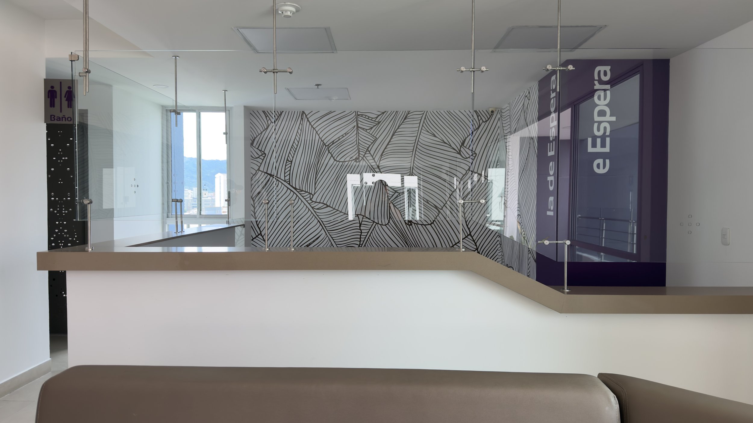 Interior view of a modern building with a glass wall, decorative black and white leaf pattern on the wall, and cityscape visible through the window. There are signs indicating toilets and a door with the word "espeja".