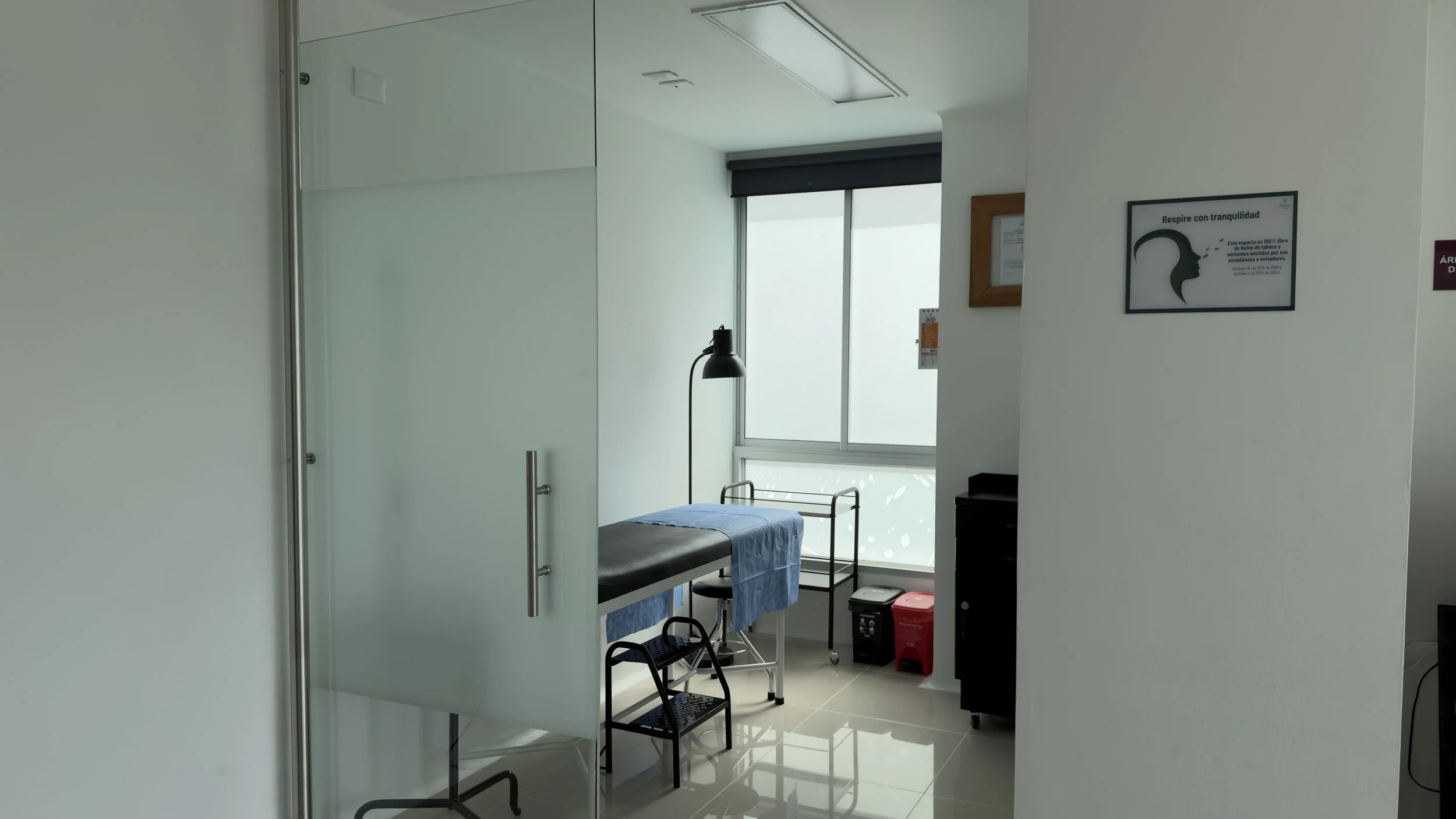 A hospital room with a patient bed covered with blue sheet, a rolling table, and a window with blinds, seen through a door with a glass panel.