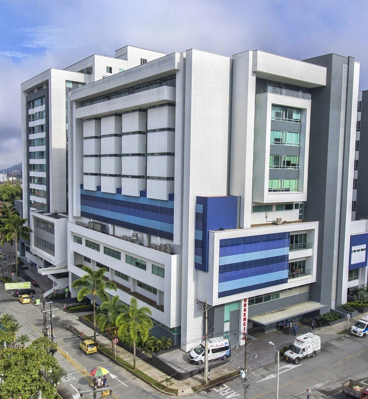 Multi-story modern white and gray building with blue accents, glass windows, and a parking lot with cars and palm trees in front.