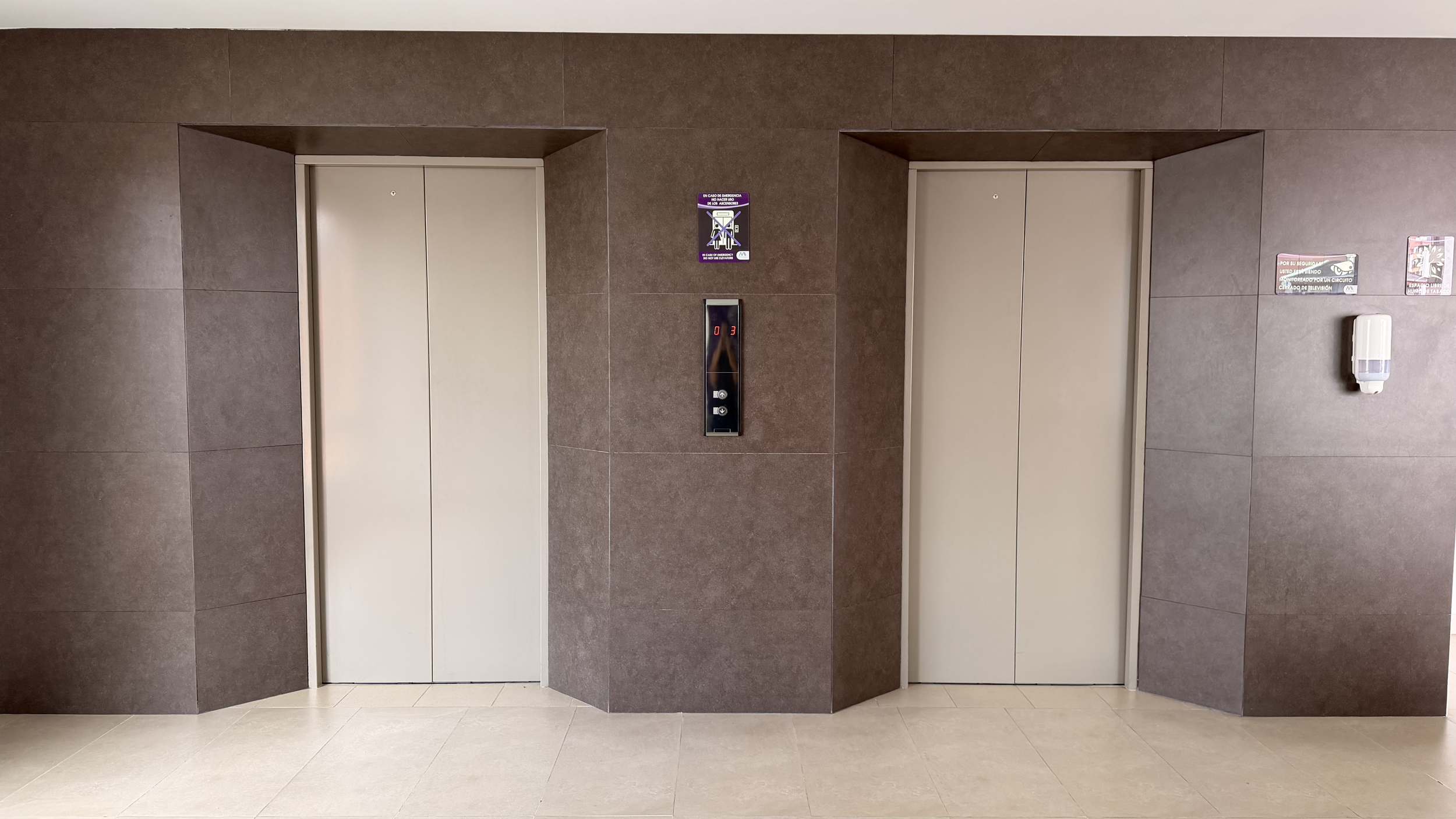 Two elevator doors in a brown tiled wall with a digital floor indicator and safety signs.