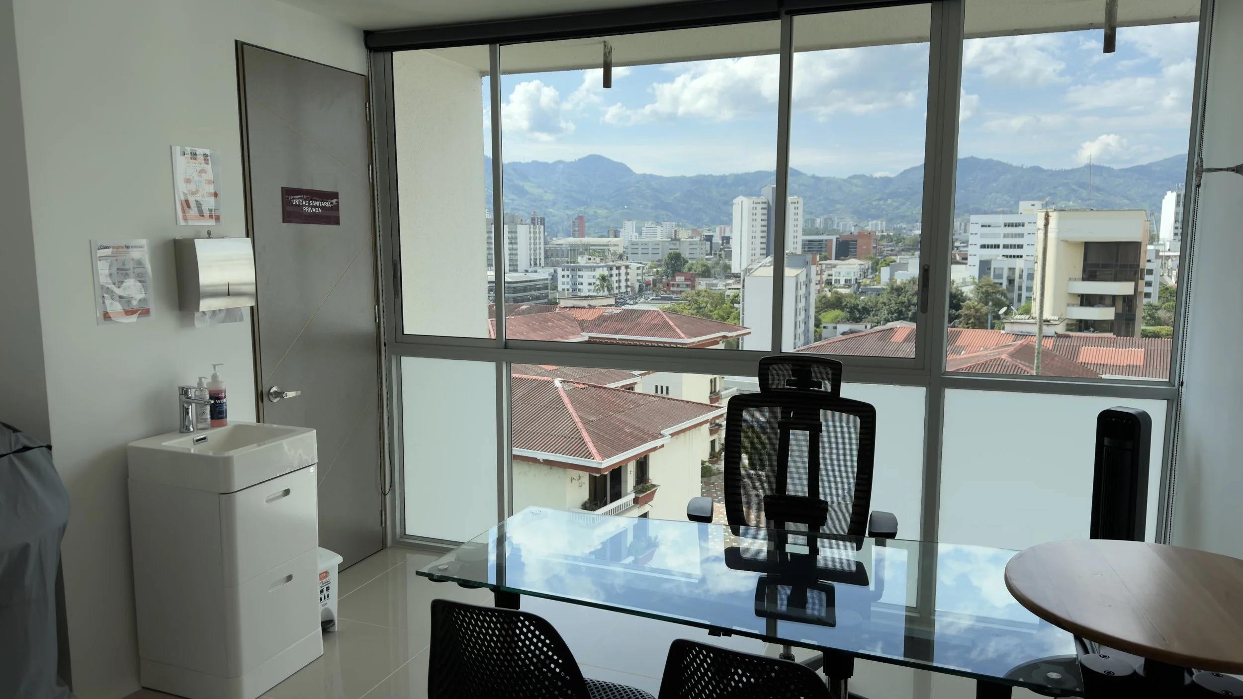 View of a city skyline with mountains in the background, taken from an office with large windows, a glass table, and office chairs.