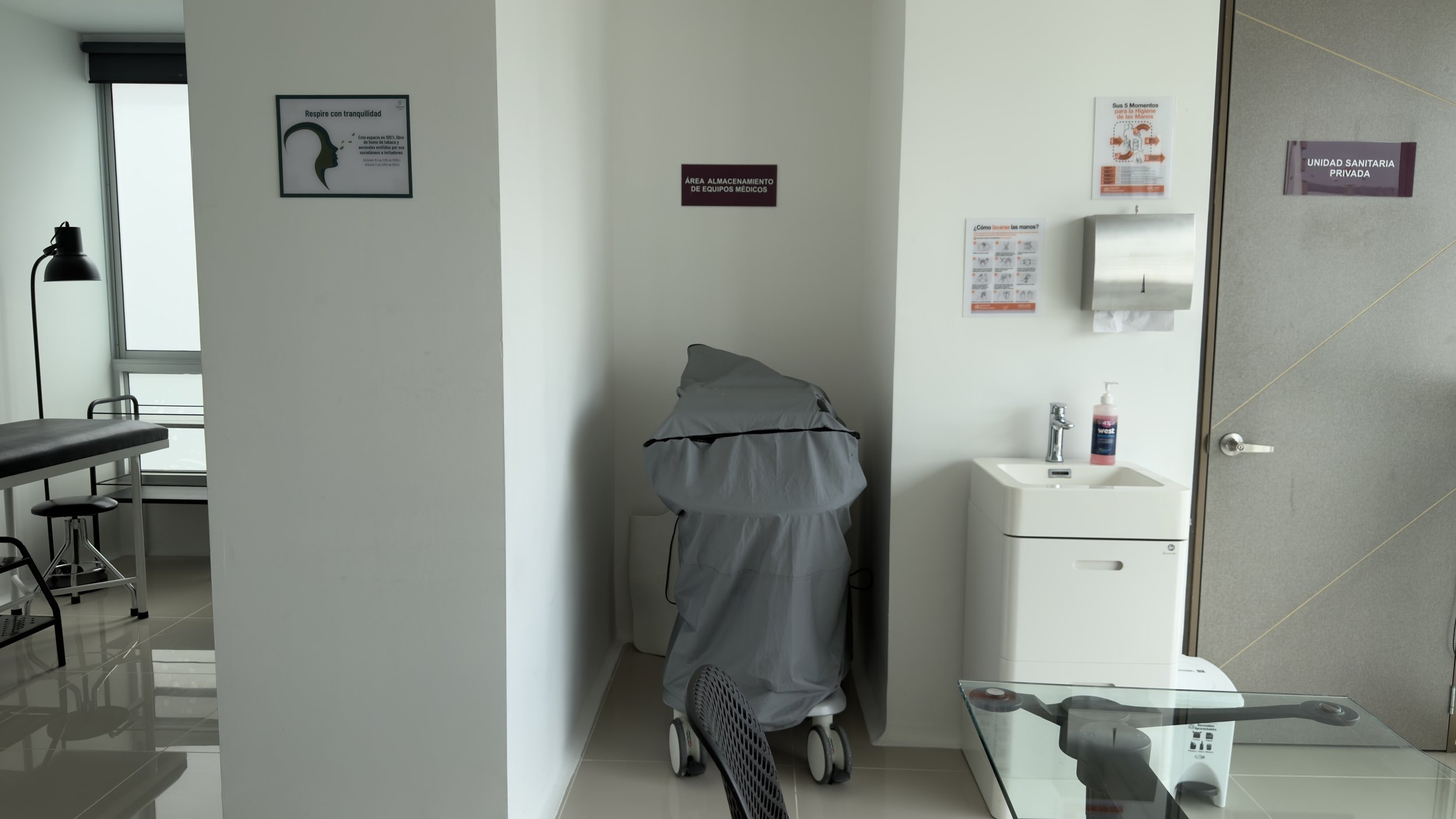 Medical storage area with a covered medical cart, a hand sanitizer pump, and warning signs on the wall, including instructions on hand hygiene and breathing exercises.