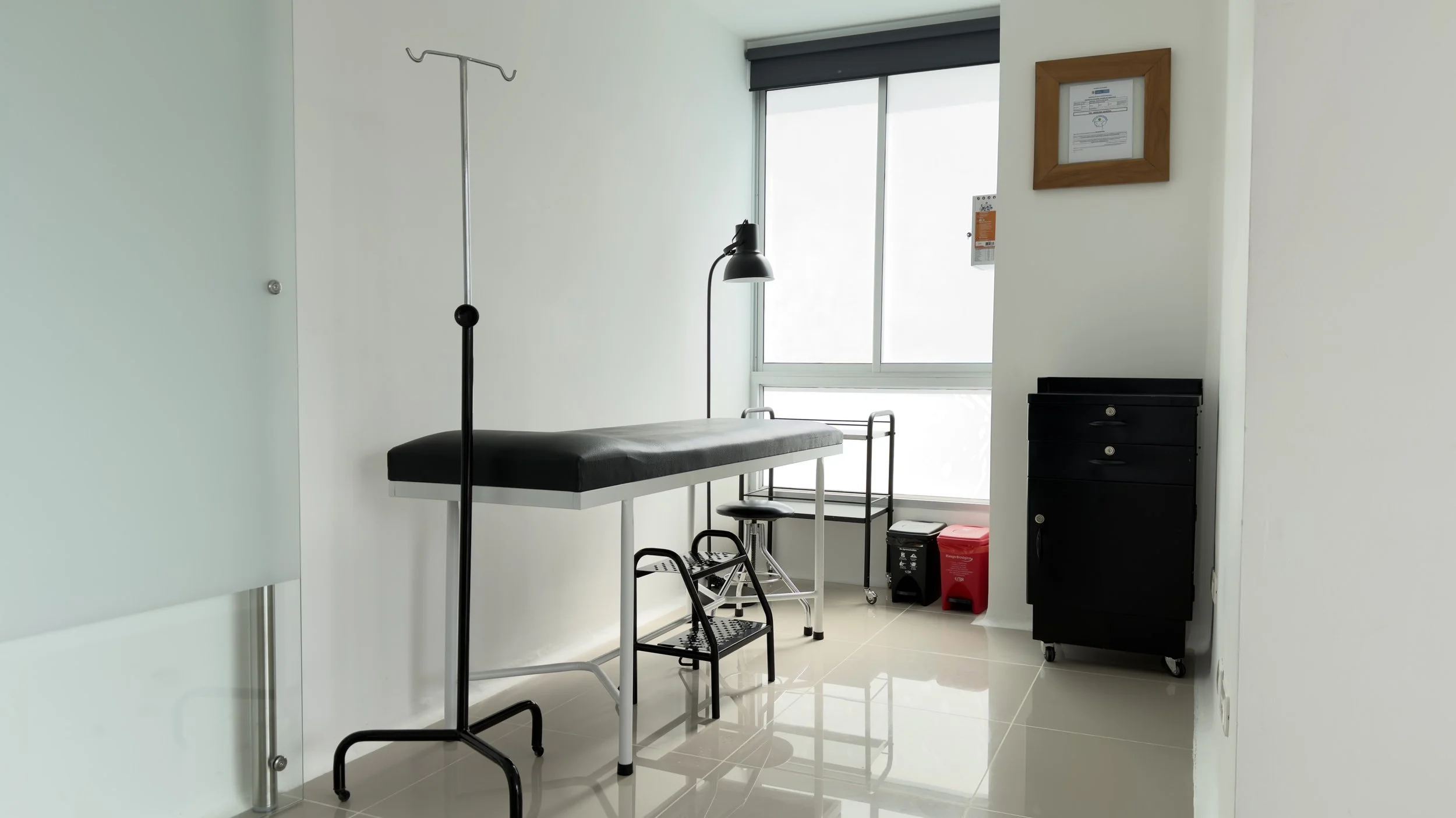 Hospital room with examination table, IV stand, chair, black cabinet, and medical equipment near large window with sunlight.