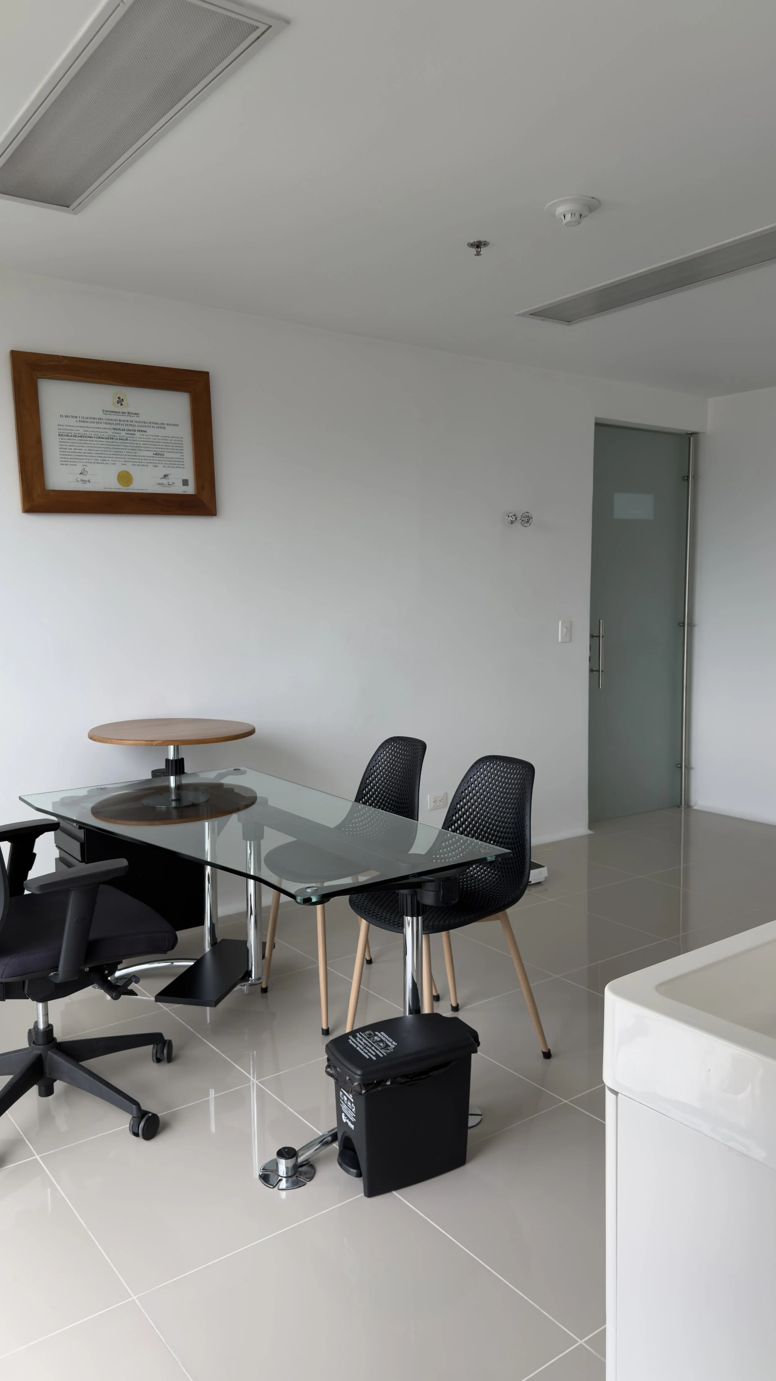 A modern conference room with a glass table, black mesh chairs, a small round wooden table, and white walls. There is a framed certificate on the wall, a trash bin, and a door in the background.