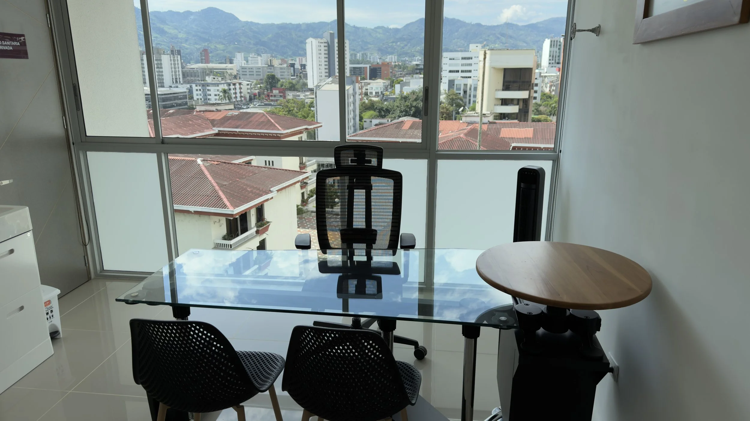 Office with glass table, black mesh office chair, round wooden table, large window overlooking city skyline and mountains.