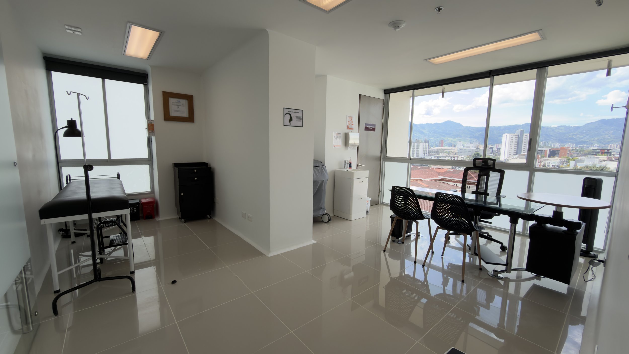 A clean, modern medical or office room with large windows showing a cityscape and mountains in the background. Features include a medical examination table, chairs, a desk with a chair, and medical equipment such as an IV stand, with bright natural l