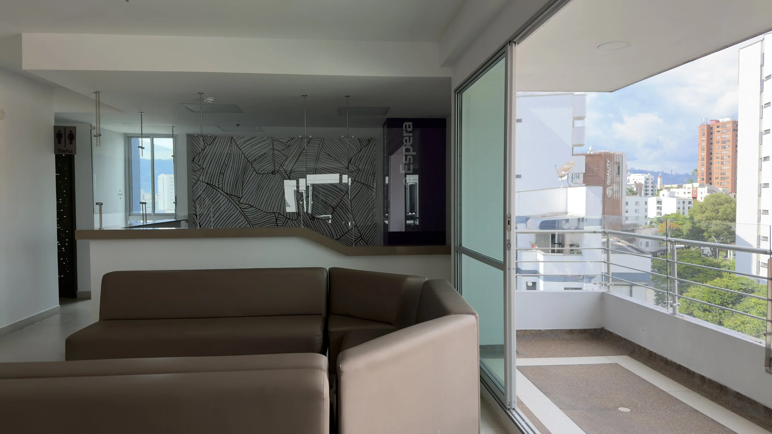 Living room with a sofa, a balcony with railing, large glass windows revealing city buildings and trees outside, and a patterned wall decor.
