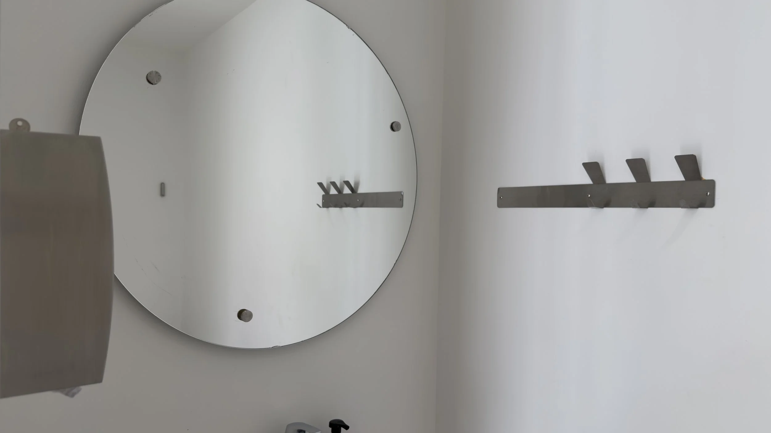 A large round mirror on the wall with a metal hook rack nearby, and a partial view of a bathroom sink with a faucet.