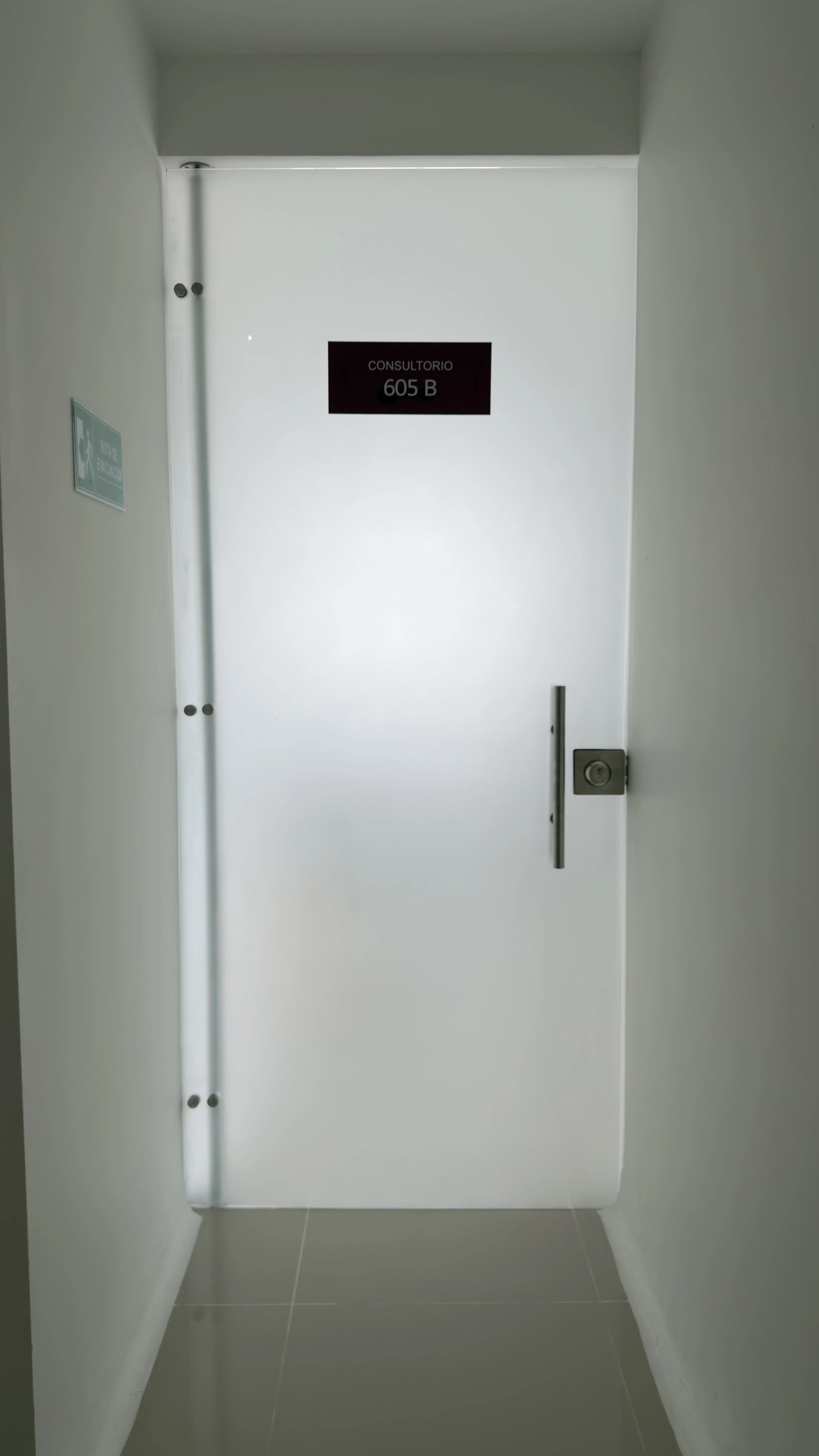 White door with a black sign that reads 'Consultorio 605 B' and a silver handle, in a white hallway with tiled floor.