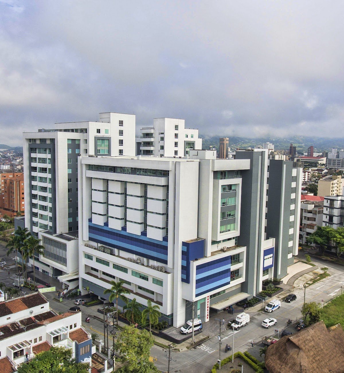 Tall modern white and gray commercial building with blue accents in an urban area, with parked cars and palm trees in the foreground, and other buildings and hills in the background under cloudy skies.