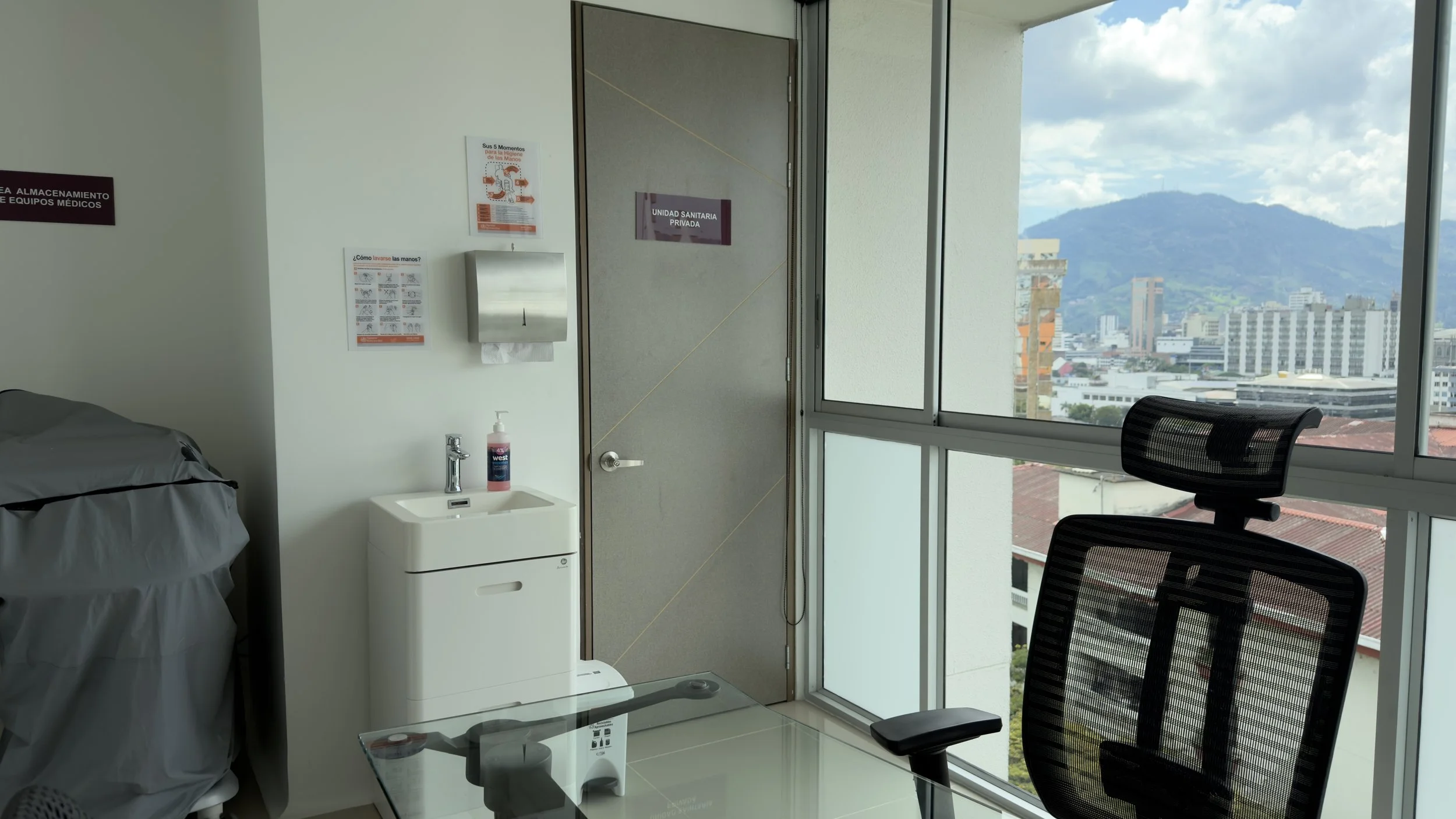 Modern clinic room with large window showing cityscape and mountains, includes a bed, a white sink with hand sanitizer, a paper towel dispenser, and an office chair.