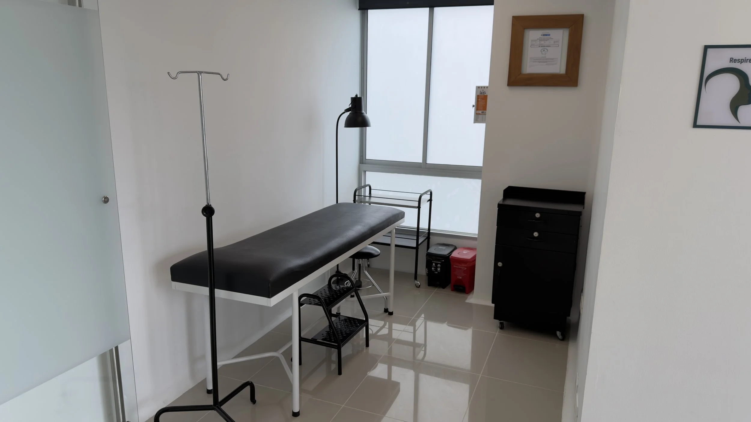 A medical examination room with a black examination table, a tall floor lamp, a small black cabinet, and medical supplies, including a red biohazard disposal container near the window.