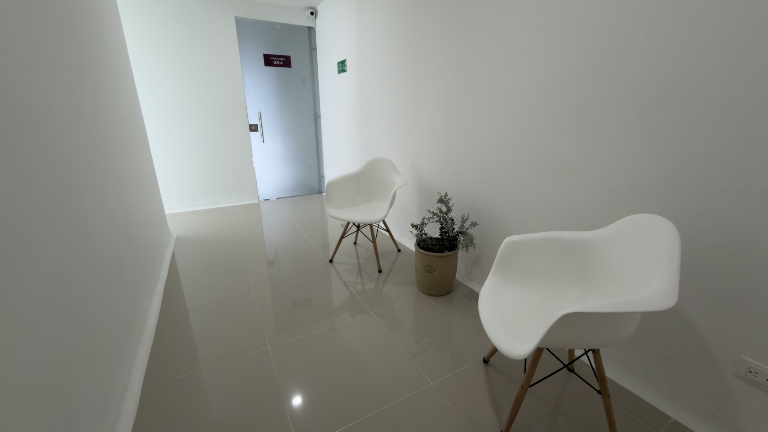 A waiting area with two white chairs and a small potted plant, with a door in the background marked 'Consultorio 605 A'.