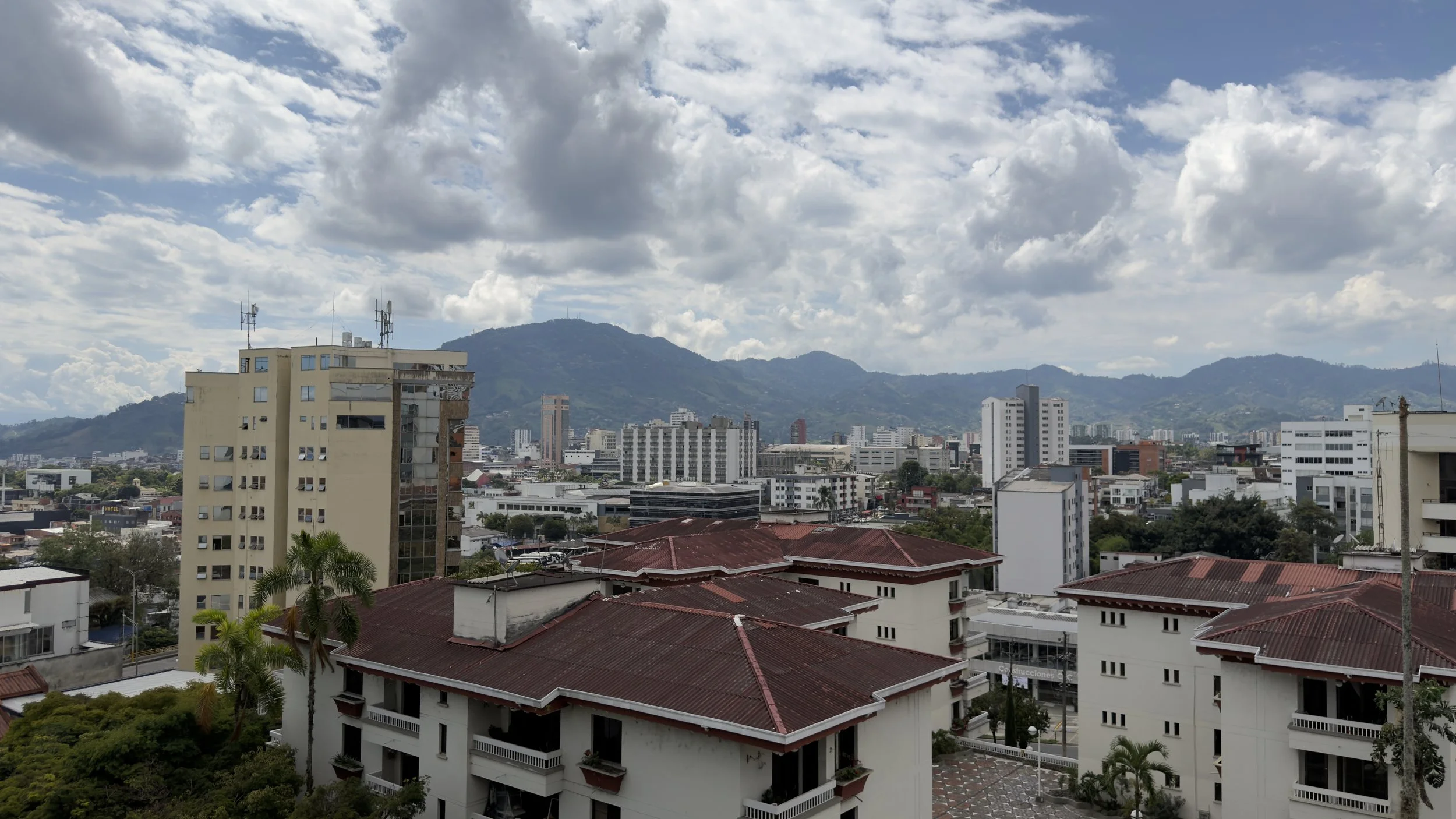 Cityscape with tall buildings, mountains in the background, and a partly cloudy sky.
