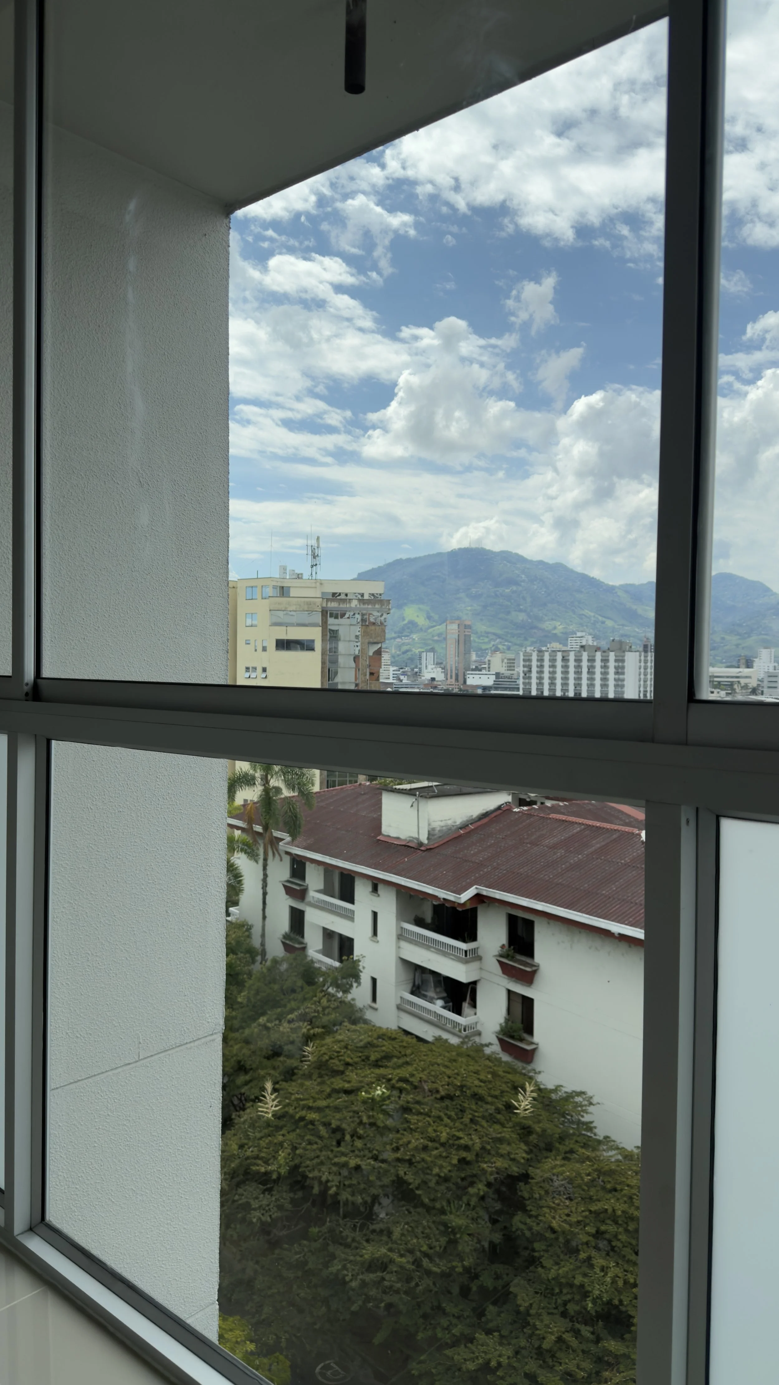 City view through a large glass window, with residential buildings, trees, and mountains in the background under a partly cloudy sky.