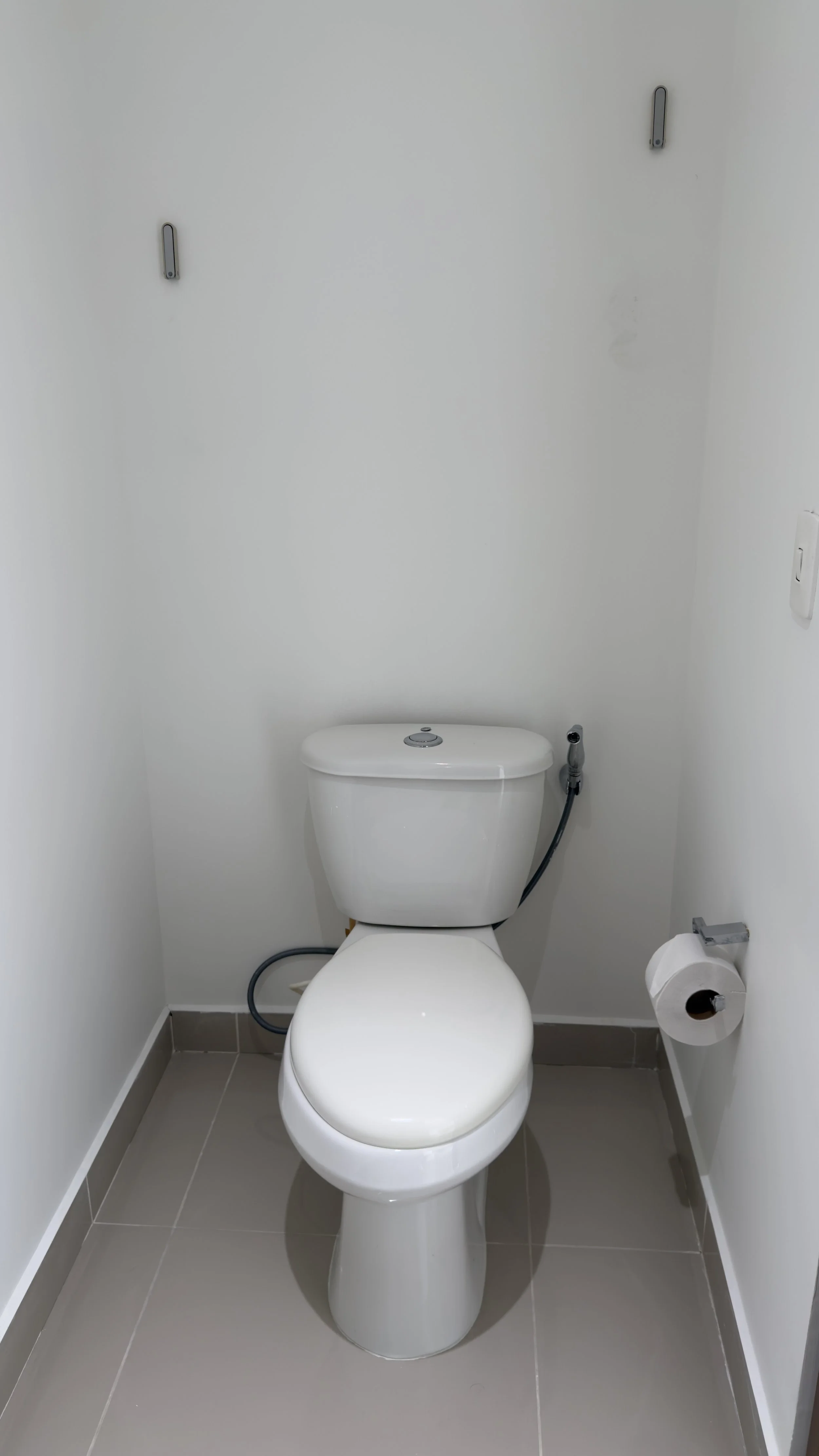 A clean white toilet with a tank and seat, situated in a small bathroom with light gray tile flooring and white walls. There is a roll of toilet paper on a holder to the right of the toilet and a toilet bidet spray attached to the side of the tank.