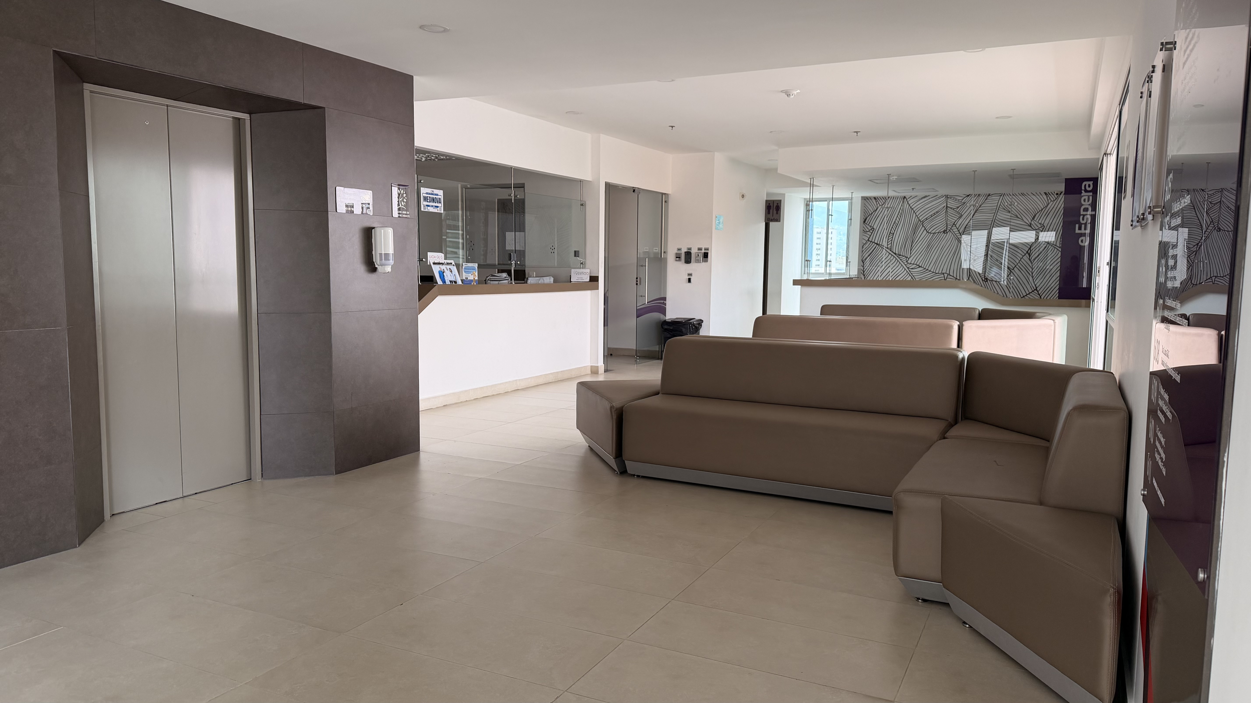 Lobby area with beige couches, elevator on the left, reception desk in the middle, large window at the back, and patterned wall art.