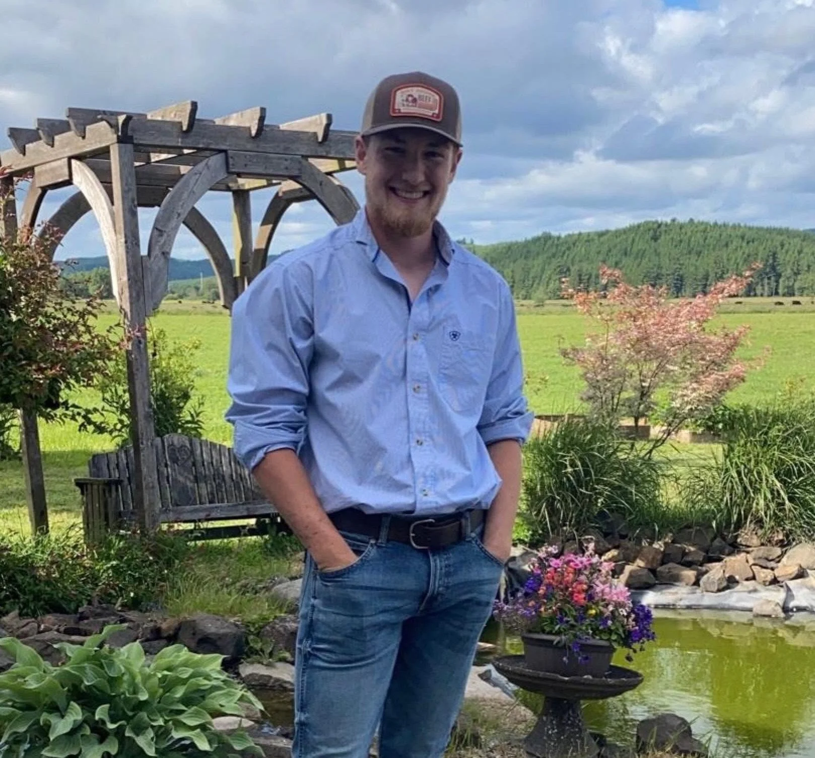 Young man in a light blue shirt and jeans standing outdoors in a garden with a pond, colorful flowers, a wooden garden structure, and rolling green hills in the background under a cloudy sky.