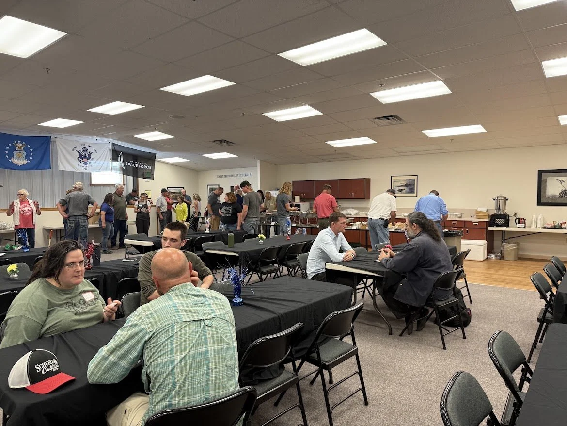 People gathered in a community hall with tables and chairs, some are eating or talking, others are standing in line at a buffet-style food service area in the background. Flags hang on the wall.