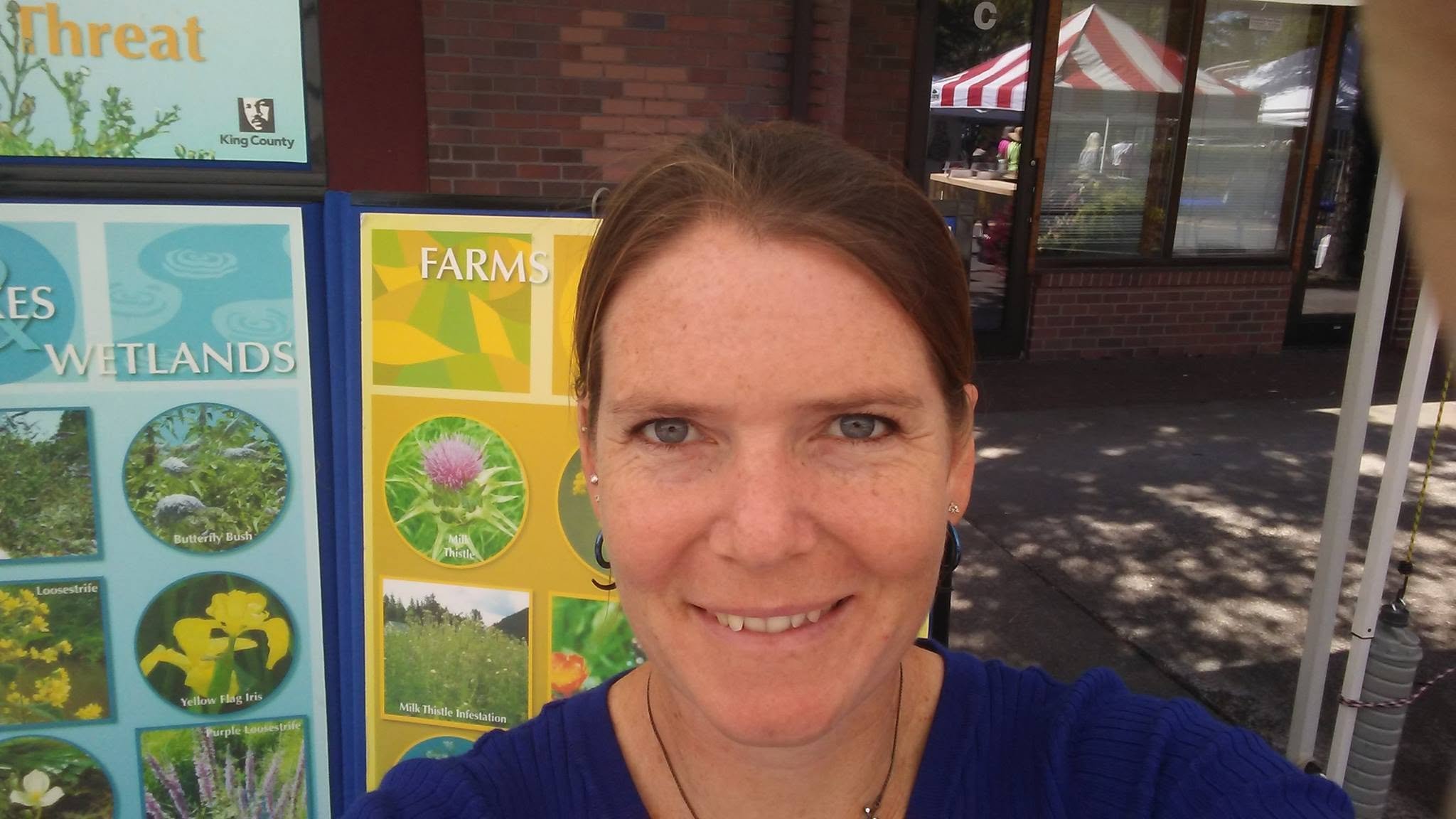 A woman taking a selfie outdoors in front of educational posters about wetlands and farms, with a brick building and tents visible in the background.