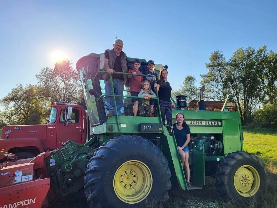 A group of people, including children and adults, are standing on and around a green John Deere tractor in a grassy field with trees in the background and the sun shining brightly in the sky.