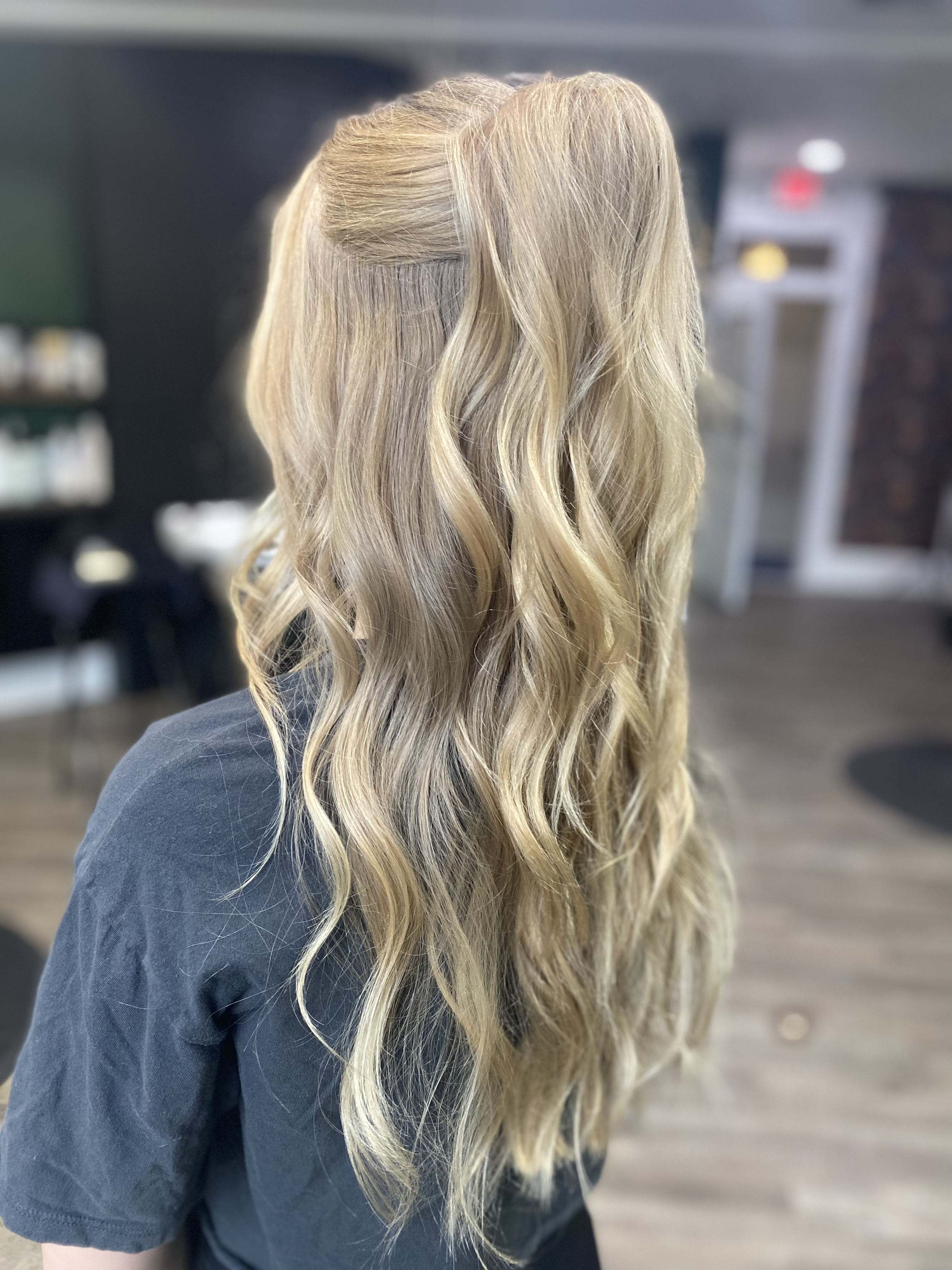 Woman with long, wavy blonde hair in a salon or studio setting, viewed from behind.