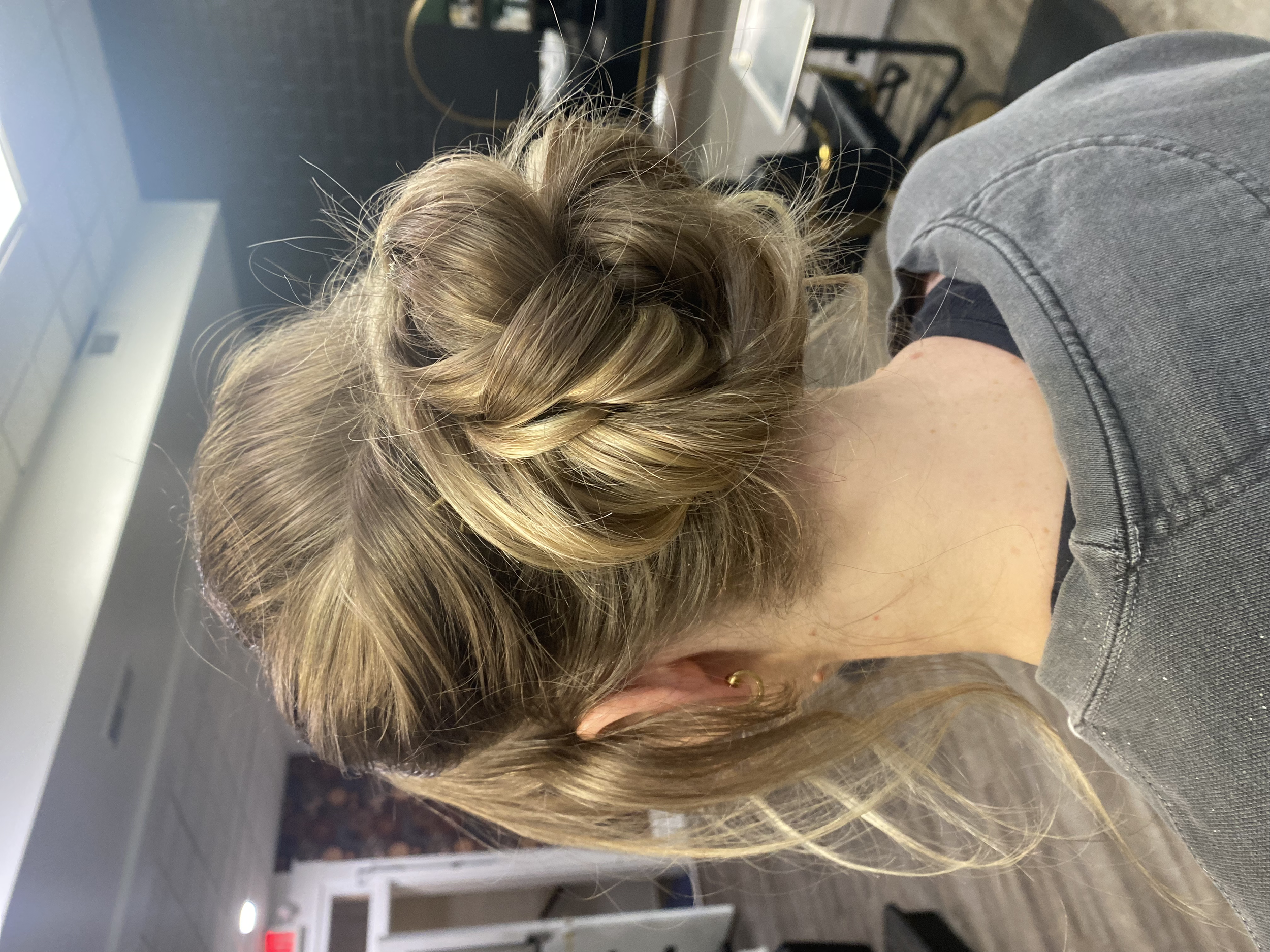 A woman with blonde hair styled in an elegant updo, in what appears to be a salon. She is wearing a grey shirt and gold hoop earrings. The background shows a salon setting with chairs, a mirror, and various salon tools.