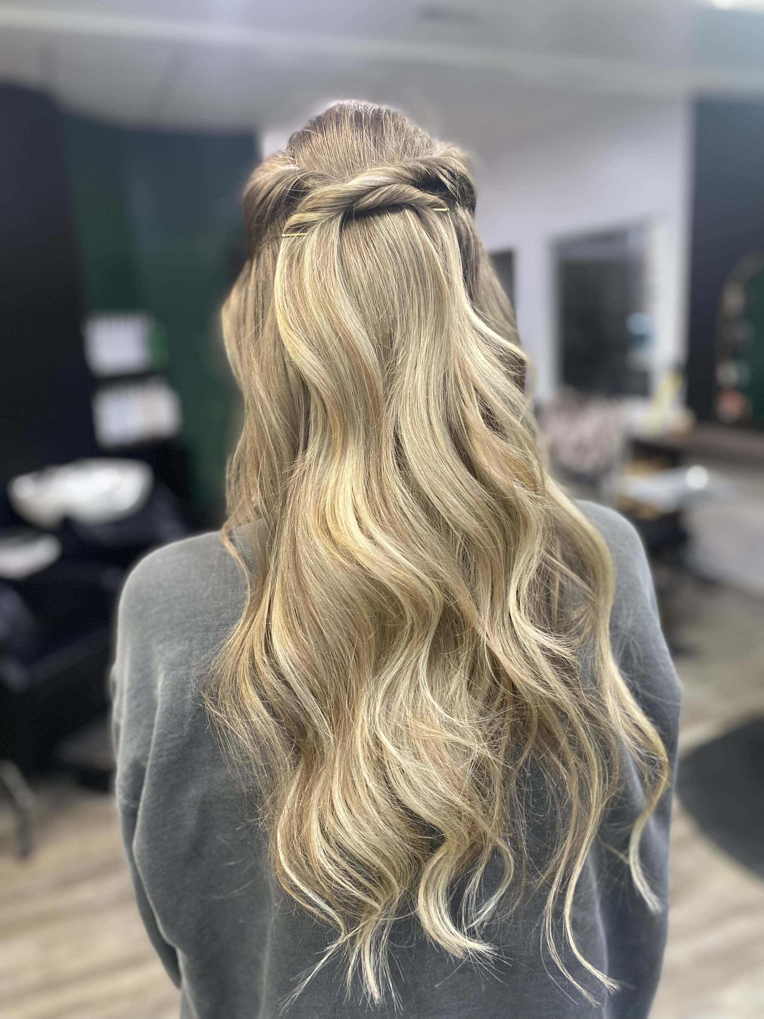 Back view of a woman with long, wavy blonde hair styled with a braid crown, wearing a gray top, standing indoors.