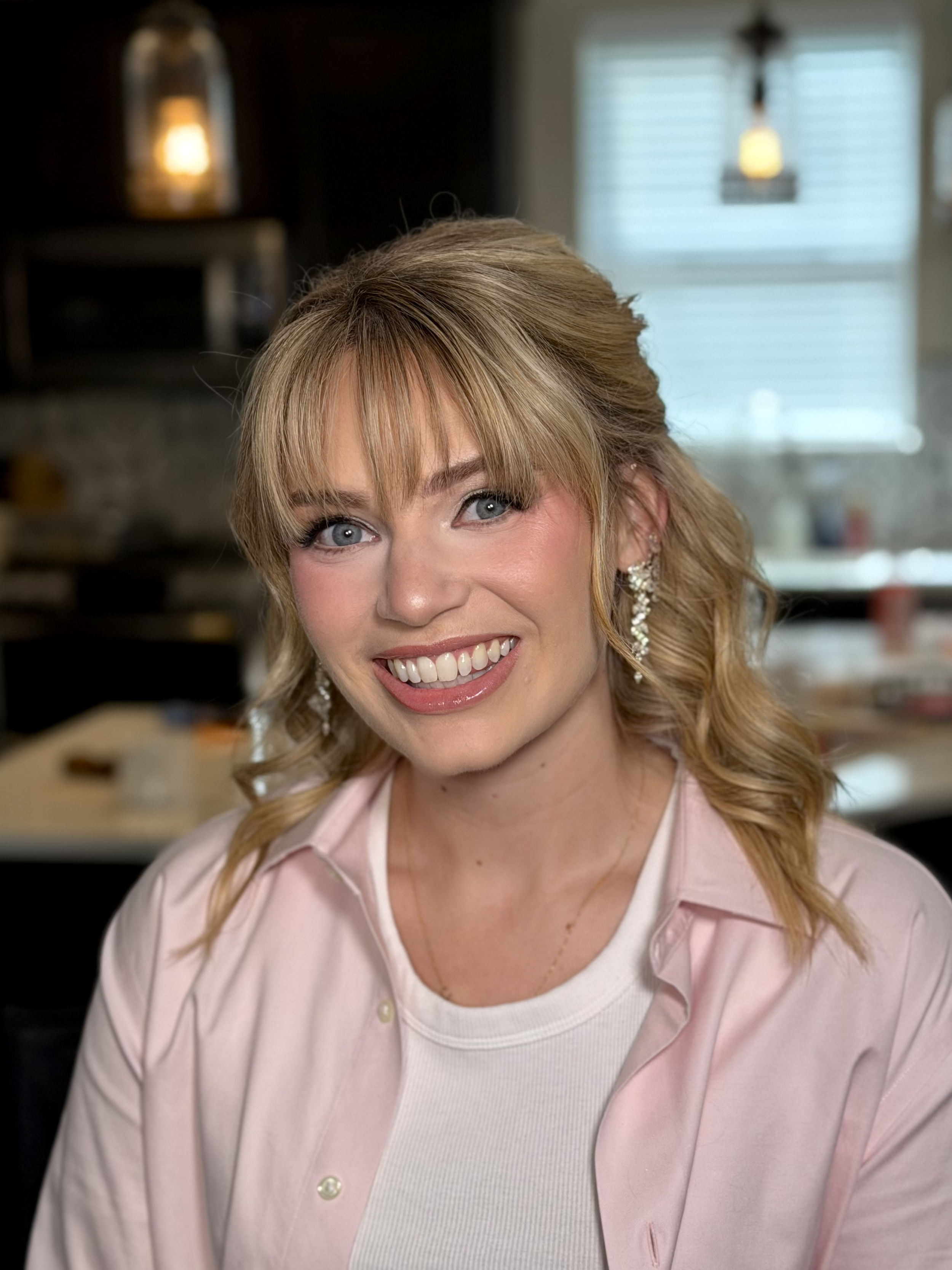 A woman with blonde hair, blue eyes, and a bright smile, wearing earrings and a light pink shirt, is smiling at the camera in a home setting.