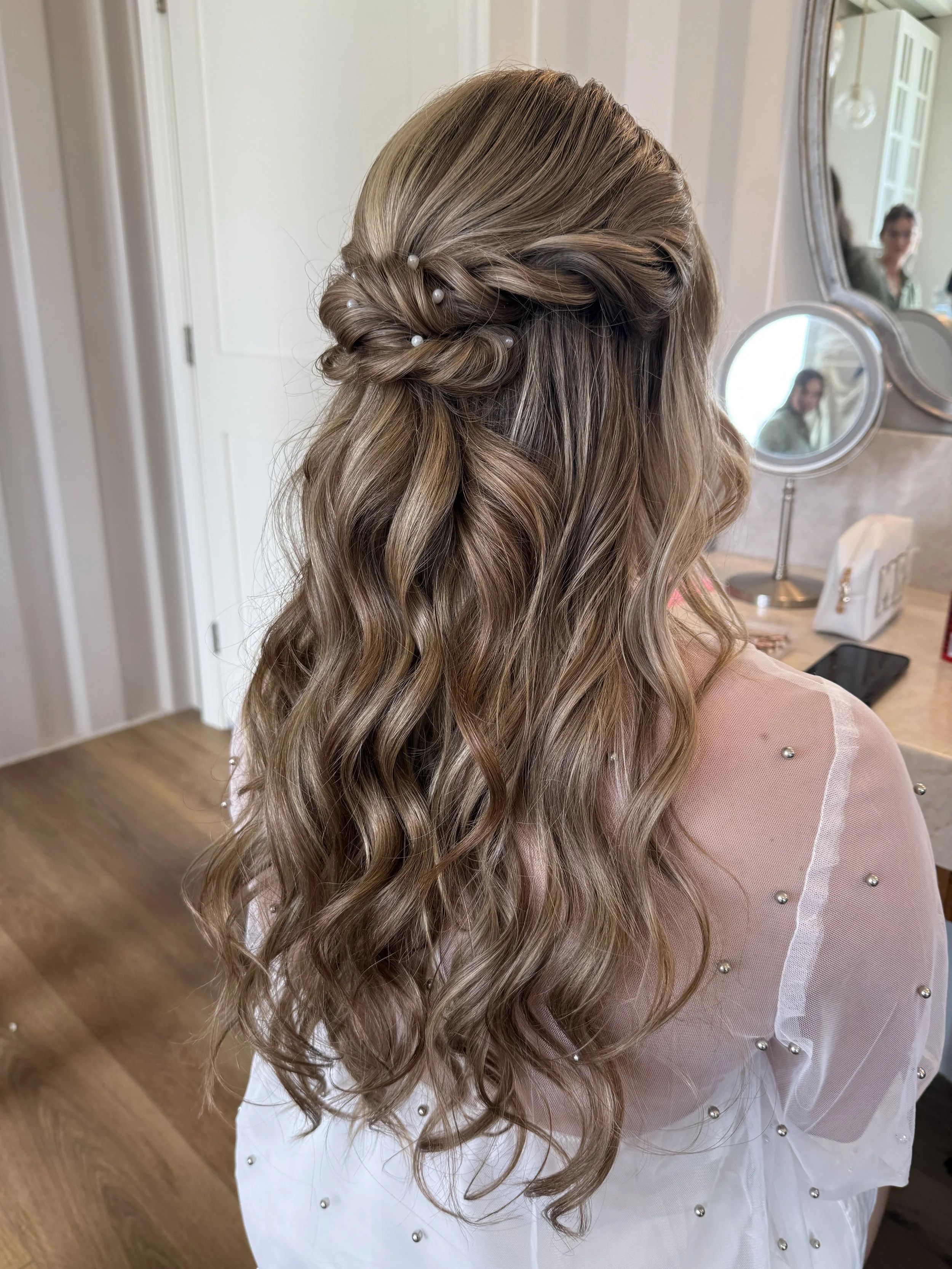 Back view of a woman with long, wavy blonde hair styled in loose curls, adorned with small white pearls. She is sitting in front of a mirror, wearing a sheer, light pink garment with pearl embellishments on the sleeves. Reflection of her face is visi