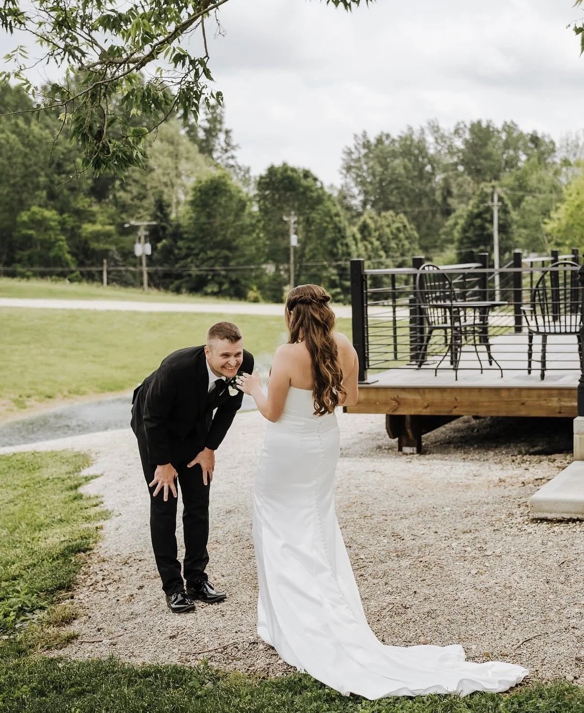 A bride with long brown hair in a white wedding gown, laughing and touching her groom's shoulder, who is in a black tuxedo, outdoors near a deck with black chairs and a table, green grass, trees, and cloudy sky.