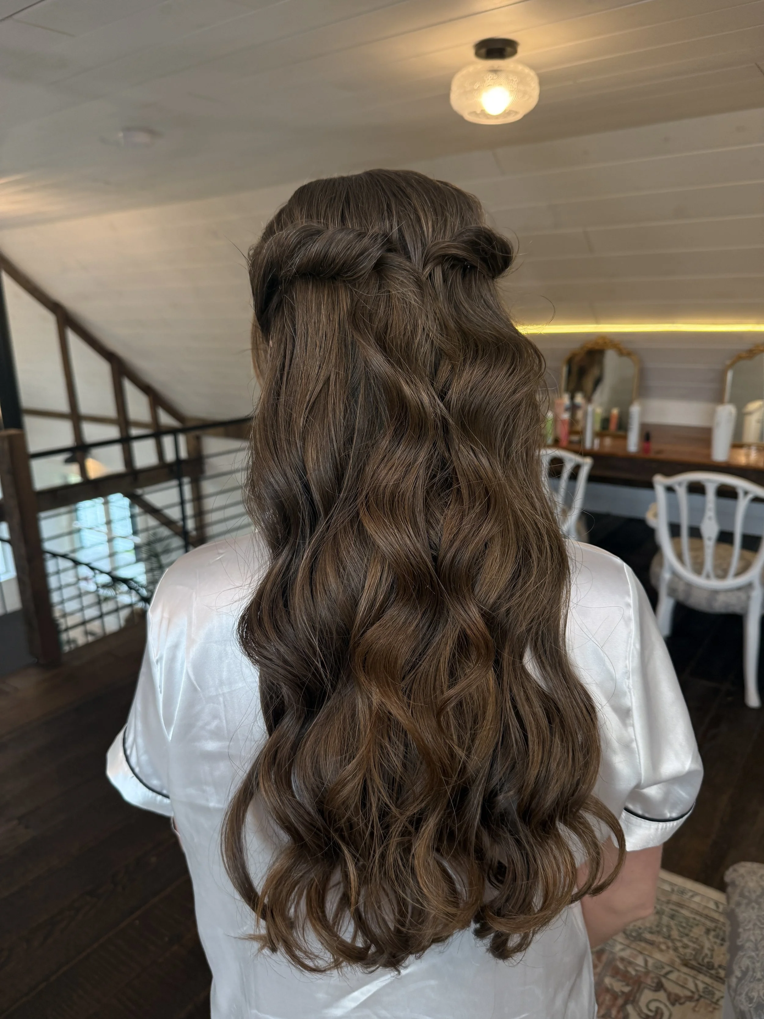 Woman with long, wavy light brown hair styled with a twisted braid Crown near the top of her head, in a room with wooden floors, white walls, and vintage-style baroque mirrors.