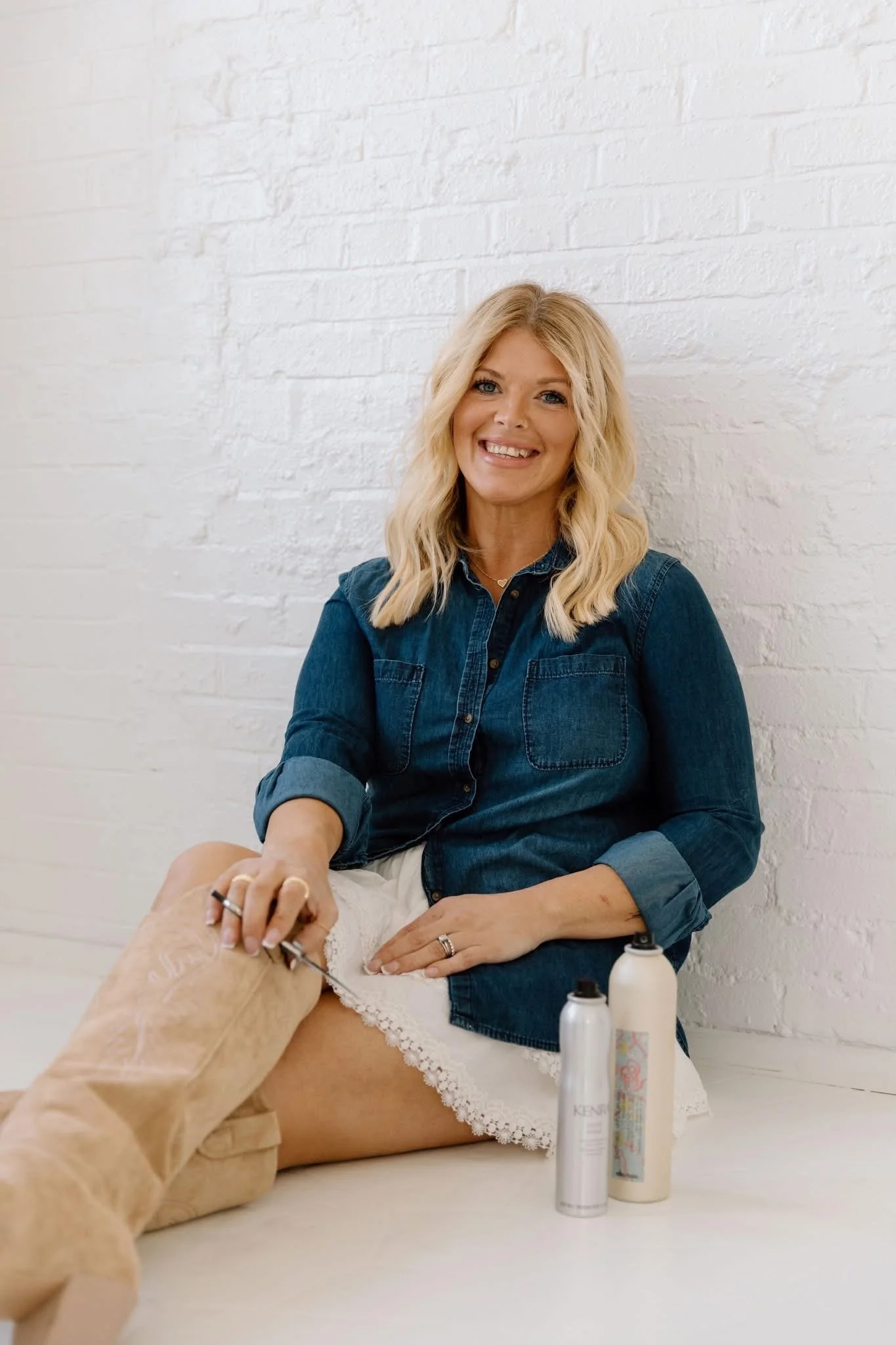 Woman smiling at the camera, sitting in front of a white brick wall, wearing a denim shirt, with skincare products in the foreground, and holding a patient's leg during a skincare or medical treatment.