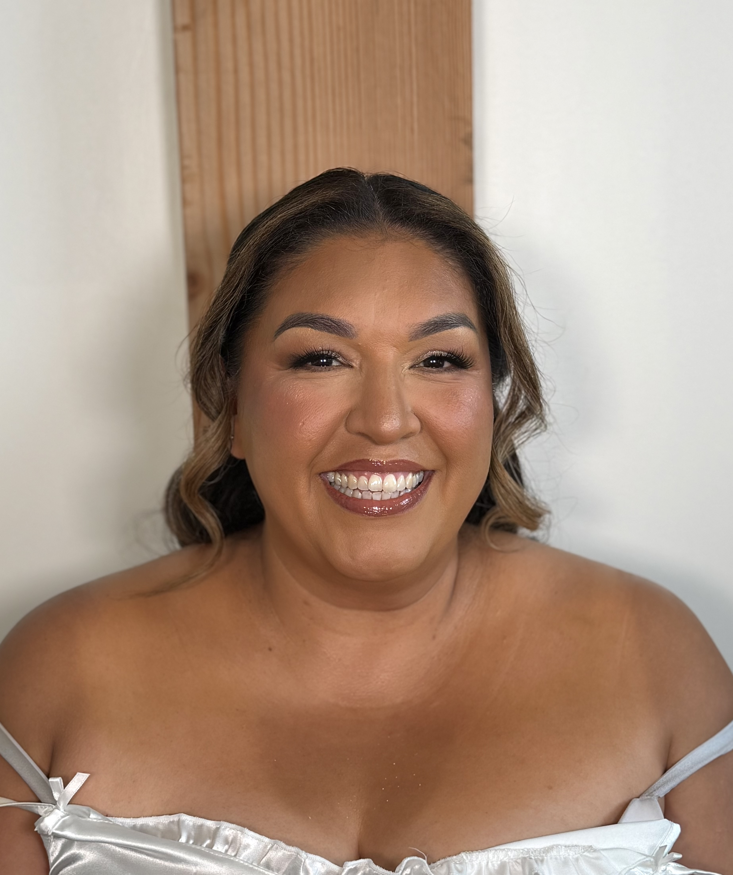 A smiling woman with wavy hair in a white satin dress with off-shoulder straps.