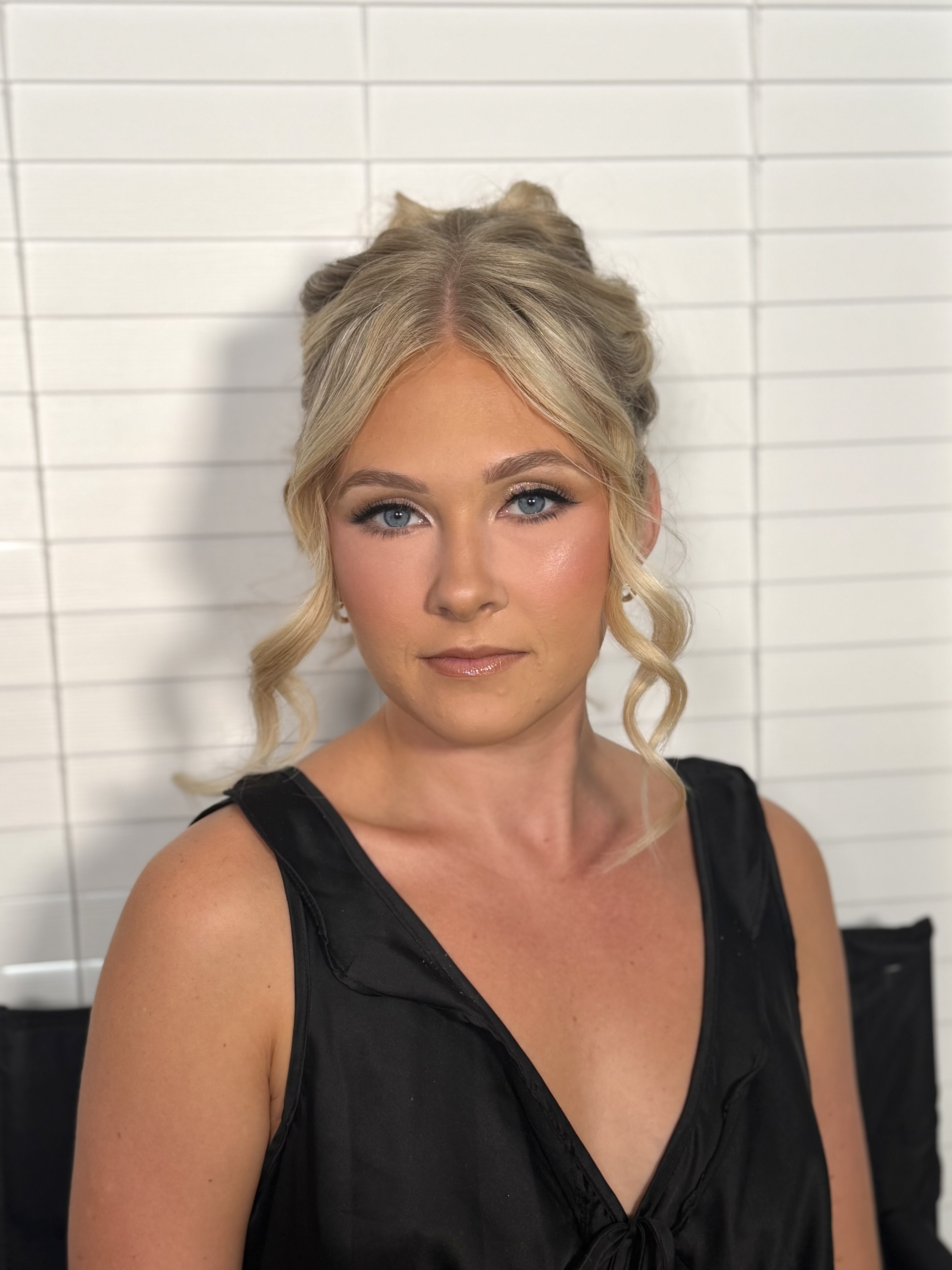 A woman with blonde hair styled in an updo with loose curls, wearing a black sleeveless dress, standing against a white tiled wall.