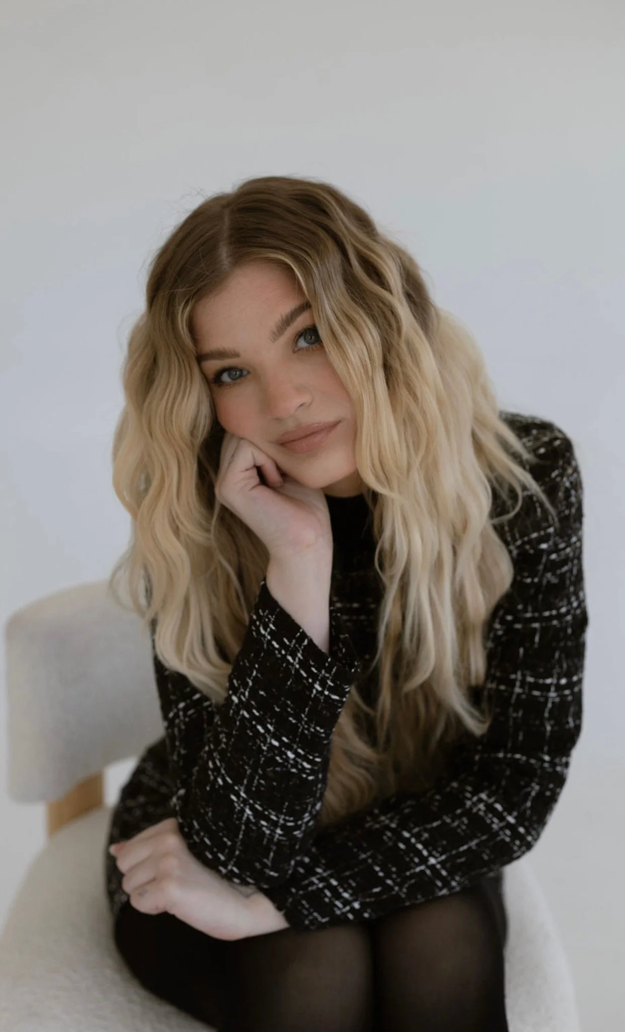 A woman with long wavy blonde hair, sitting on a beige chair with a white wall background. She is resting her chin on her hand and looking at the camera with a slight smile. She is wearing a black and white patterned long-sleeve top and black tights.